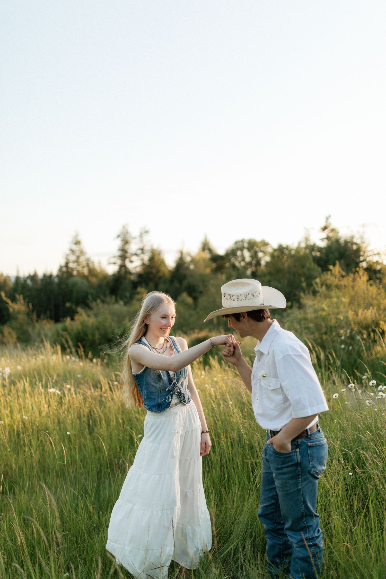 Couple in grassy field at sunset, man in cowboy hat kissing woman's hand while she smiles warmly.