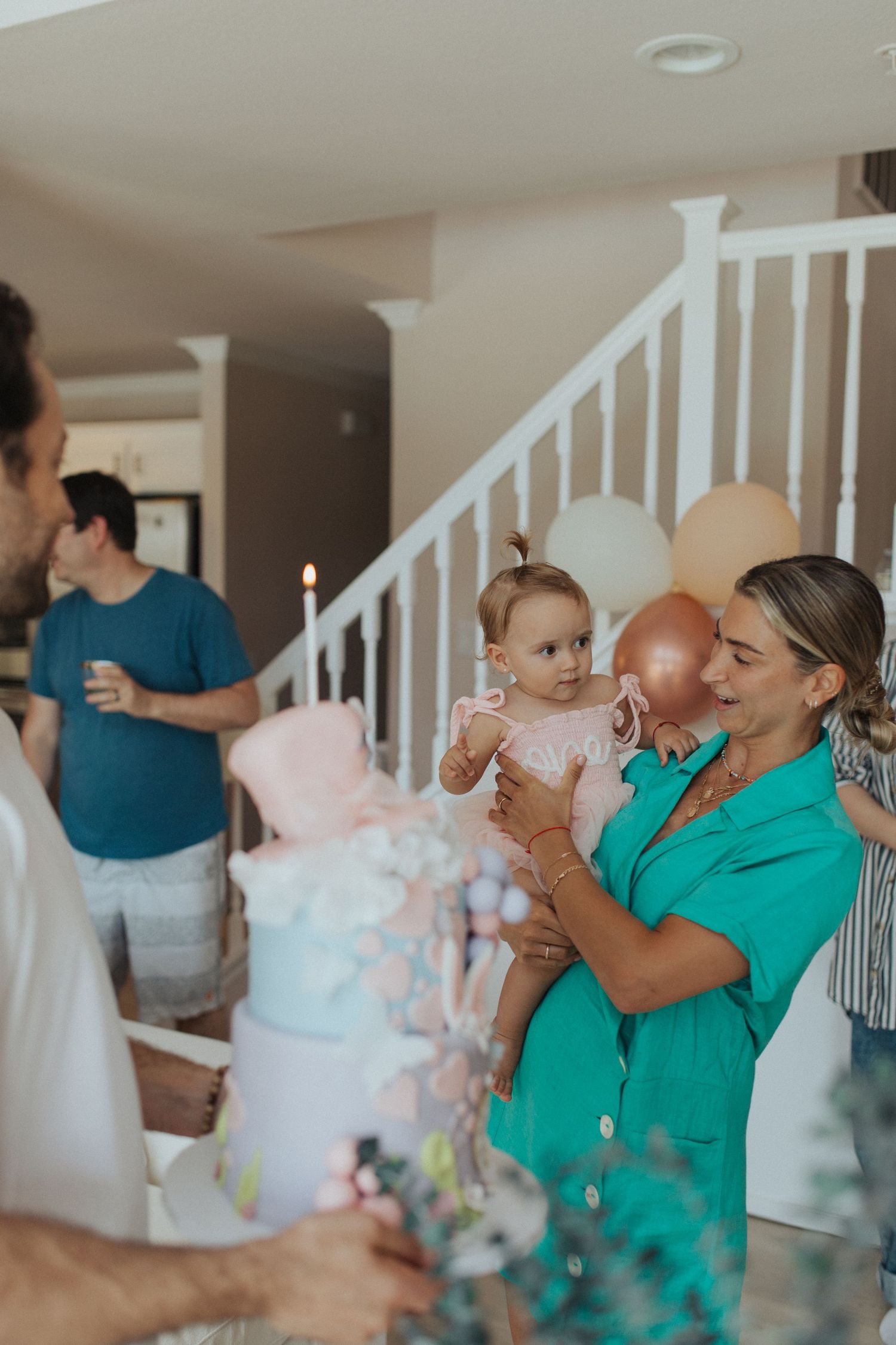 Woman in green dress holds baby girl near decorated birthday cake at indoor first birthday party.