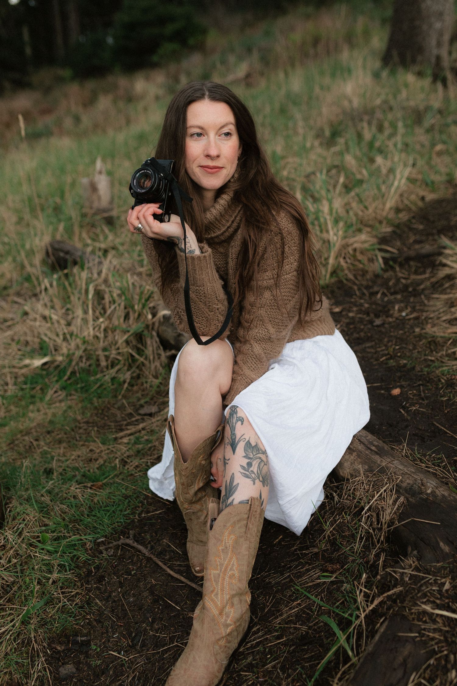 A brunette woman in a brown sweater and white skirt sits outdoors holding a vintage film camera confidently.
