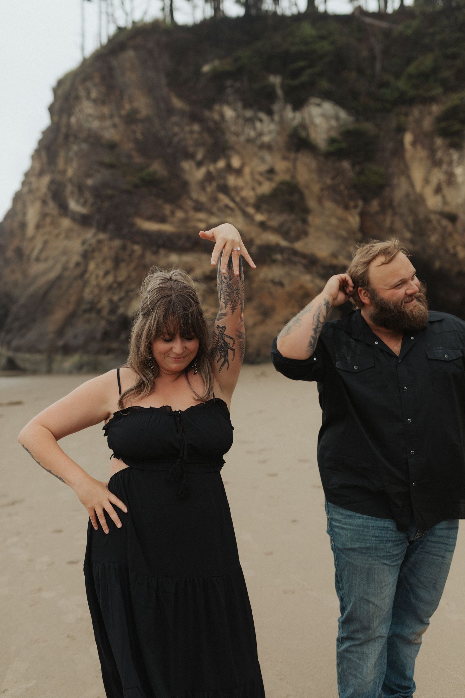Newly engaged couple walking on beach, woman showing off engagement ring with rocky cliffs behind.