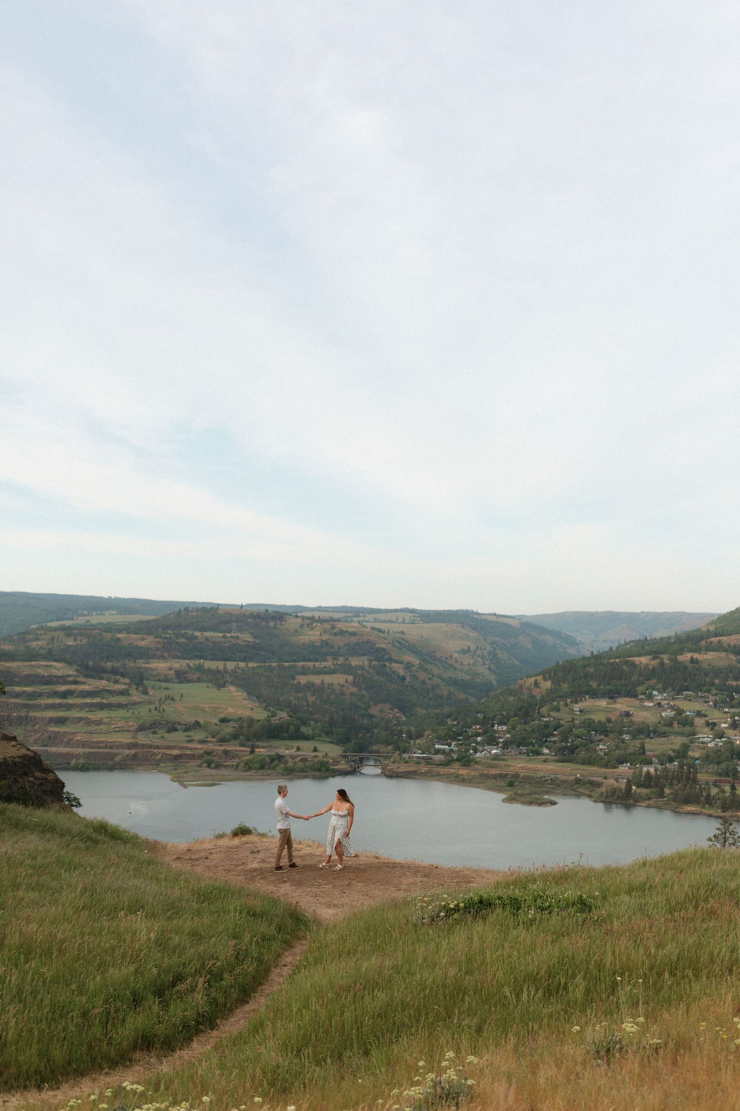Couple holding hands on a hilltop trail overlooking a serene lake and green valley below.
