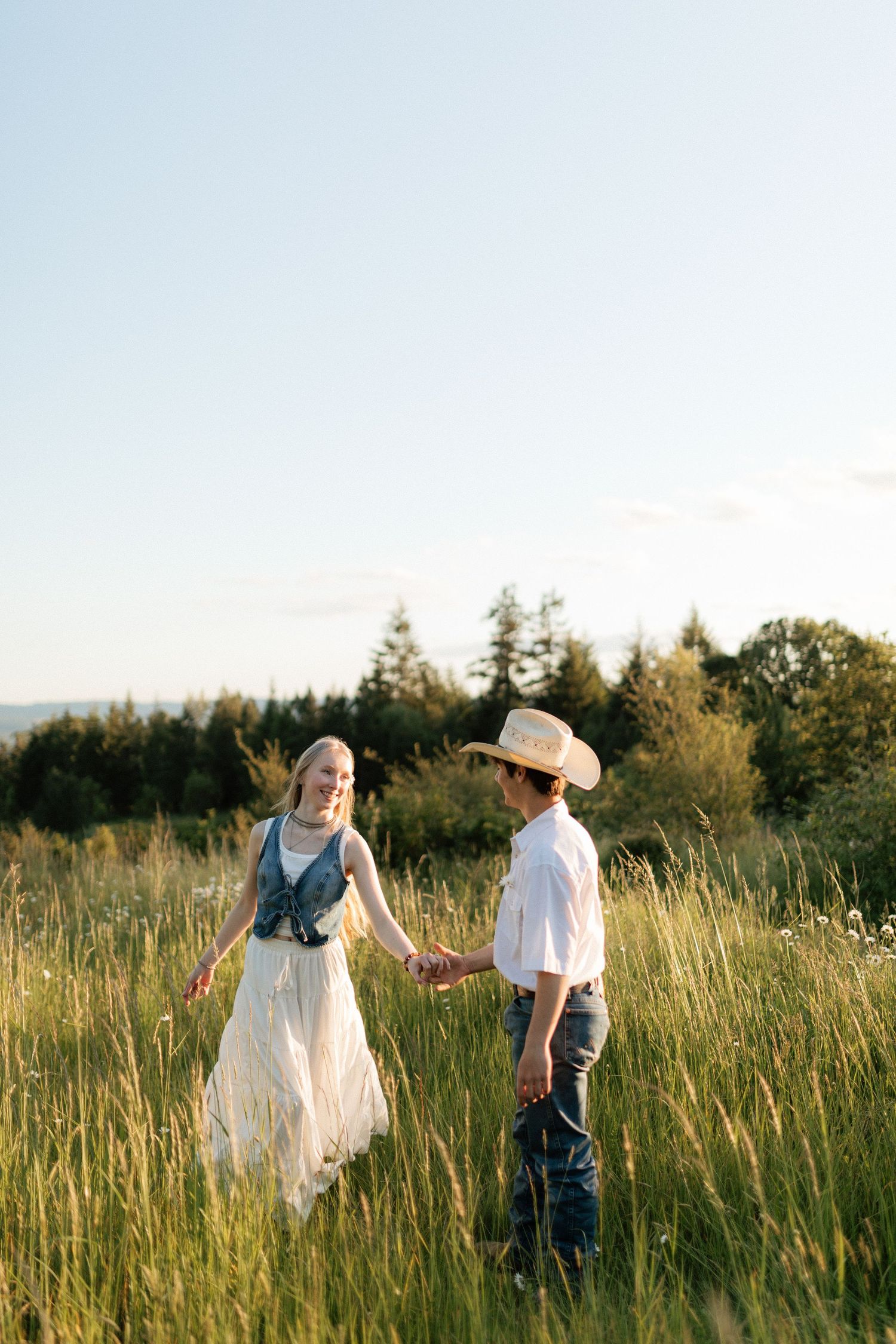 Young couple holding hands in golden meadow, woman in white dress and denim vest, man in cowboy hat.
