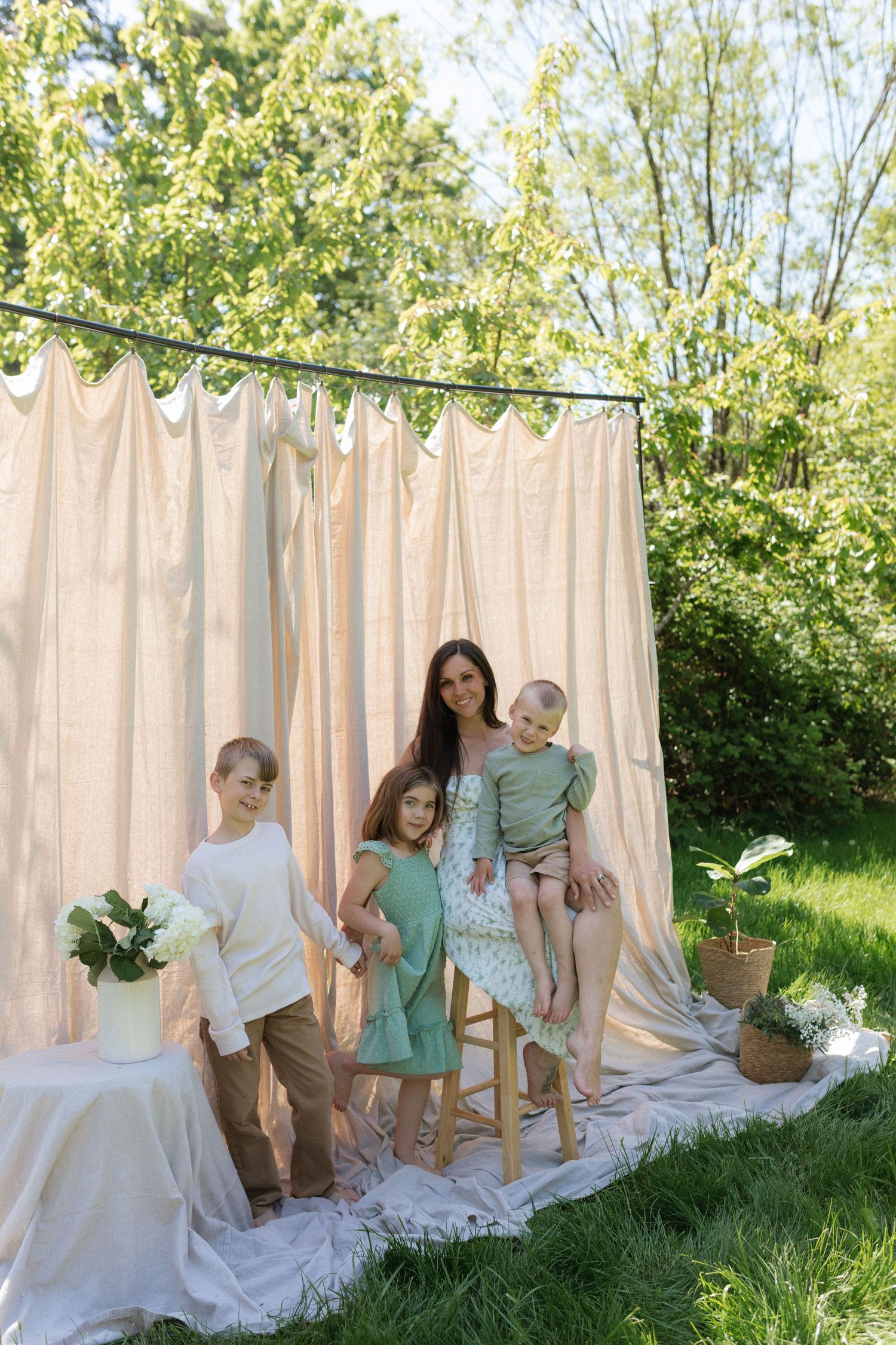 A mother poses with her three children in front of a draped backdrop in a garden.