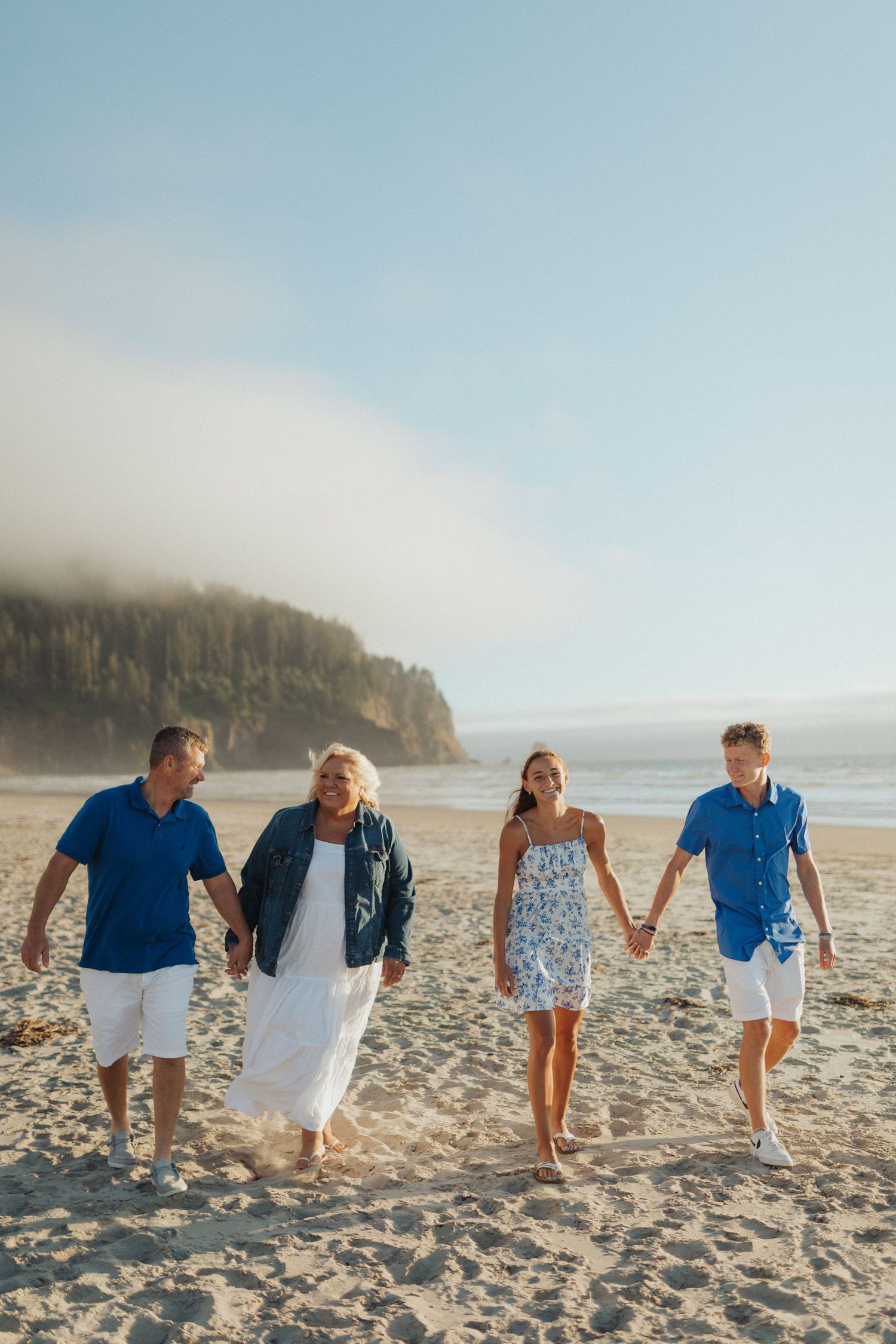 Family of four holding hands and walking together on a sunny sandy beach with foggy cliffs.