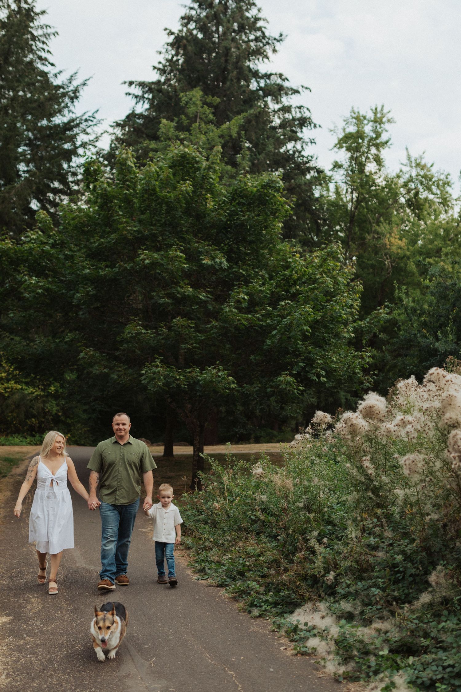 Joyful family walking their corgi dog hand in hand along a lush green park path.
