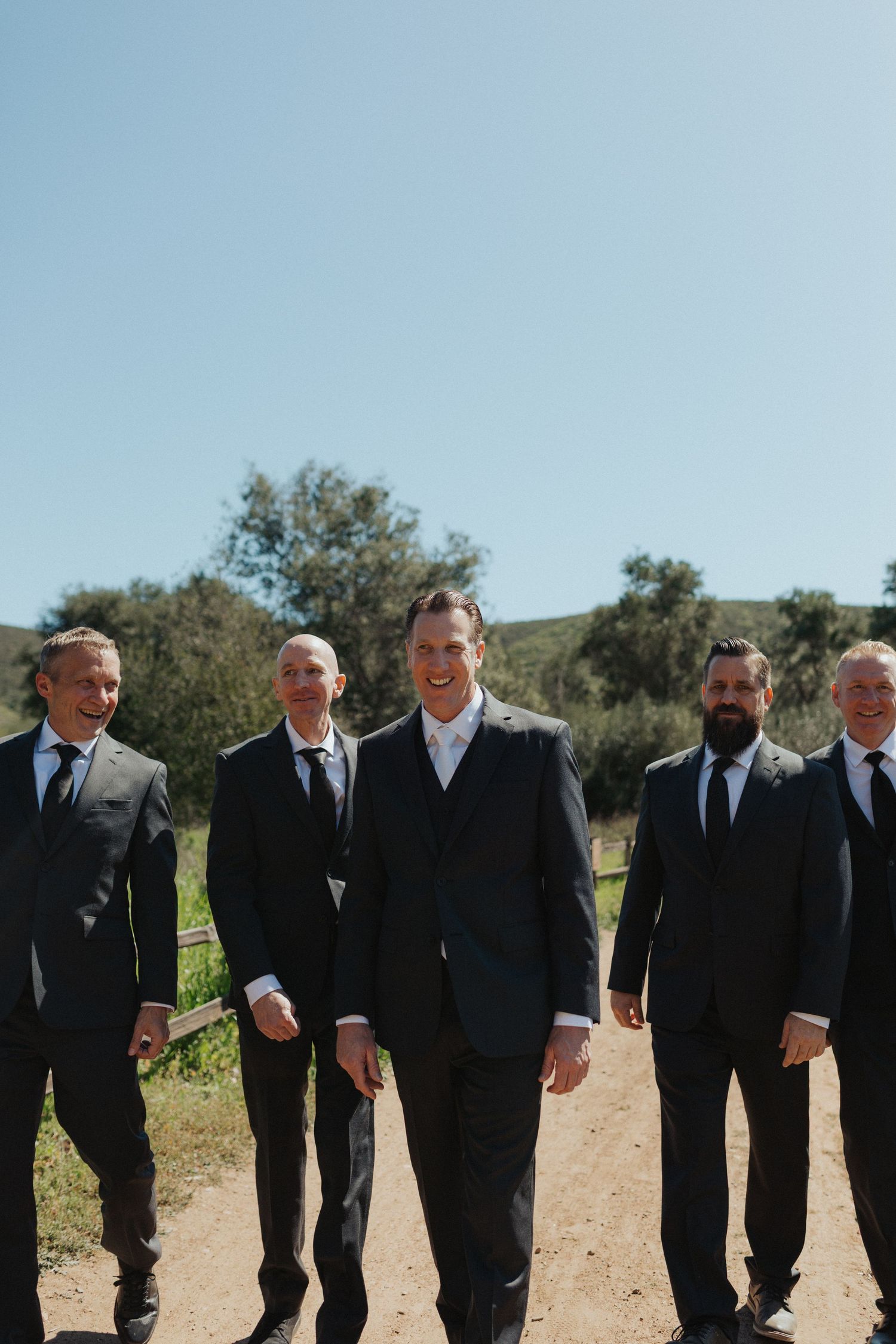 A groom and his groomsmen in matching dark suits walk confidently along a sunny dirt path with rolling hills.