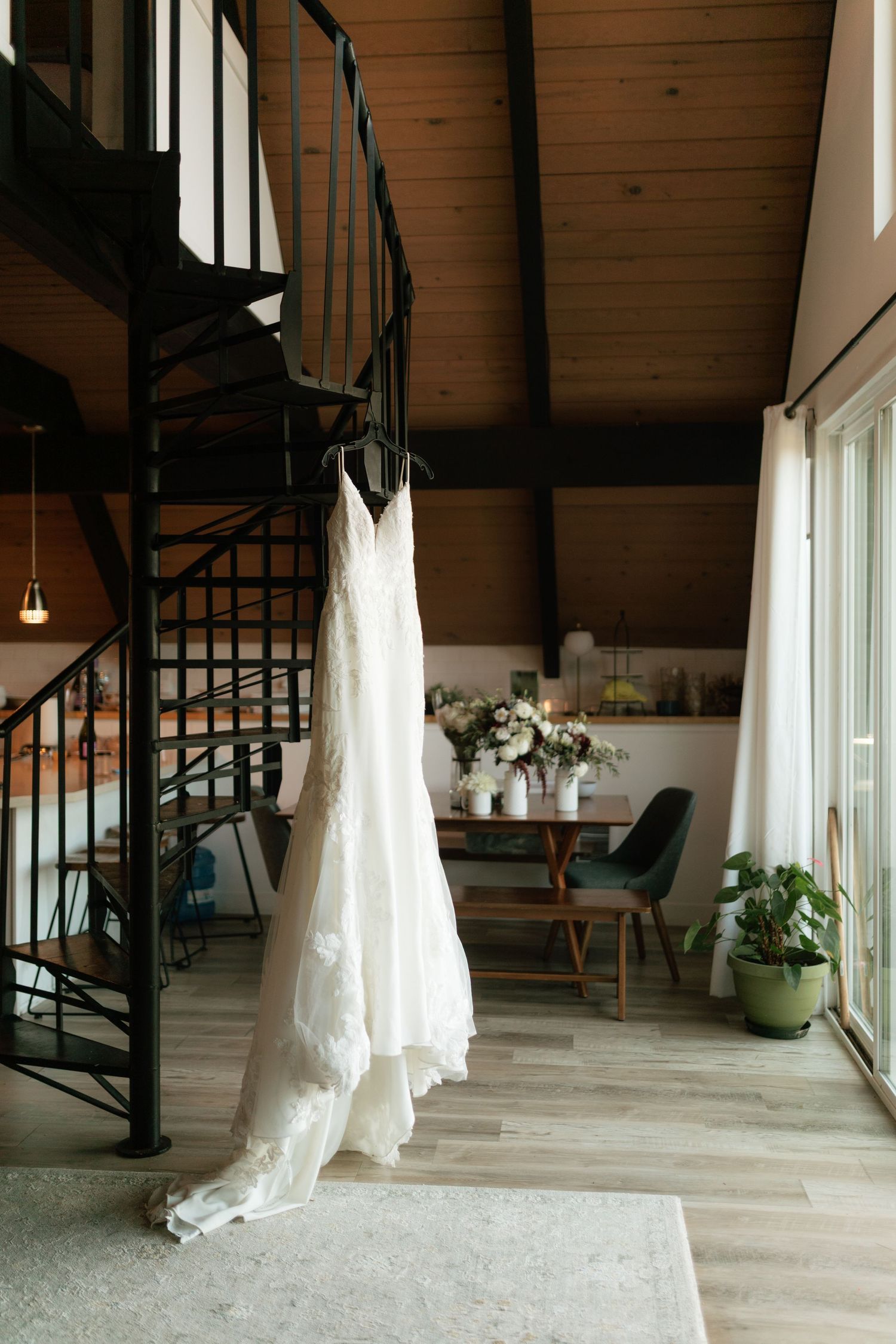 Elegant lace wedding gown hanging from a spiral staircase in a warm wooden A-frame cabin.