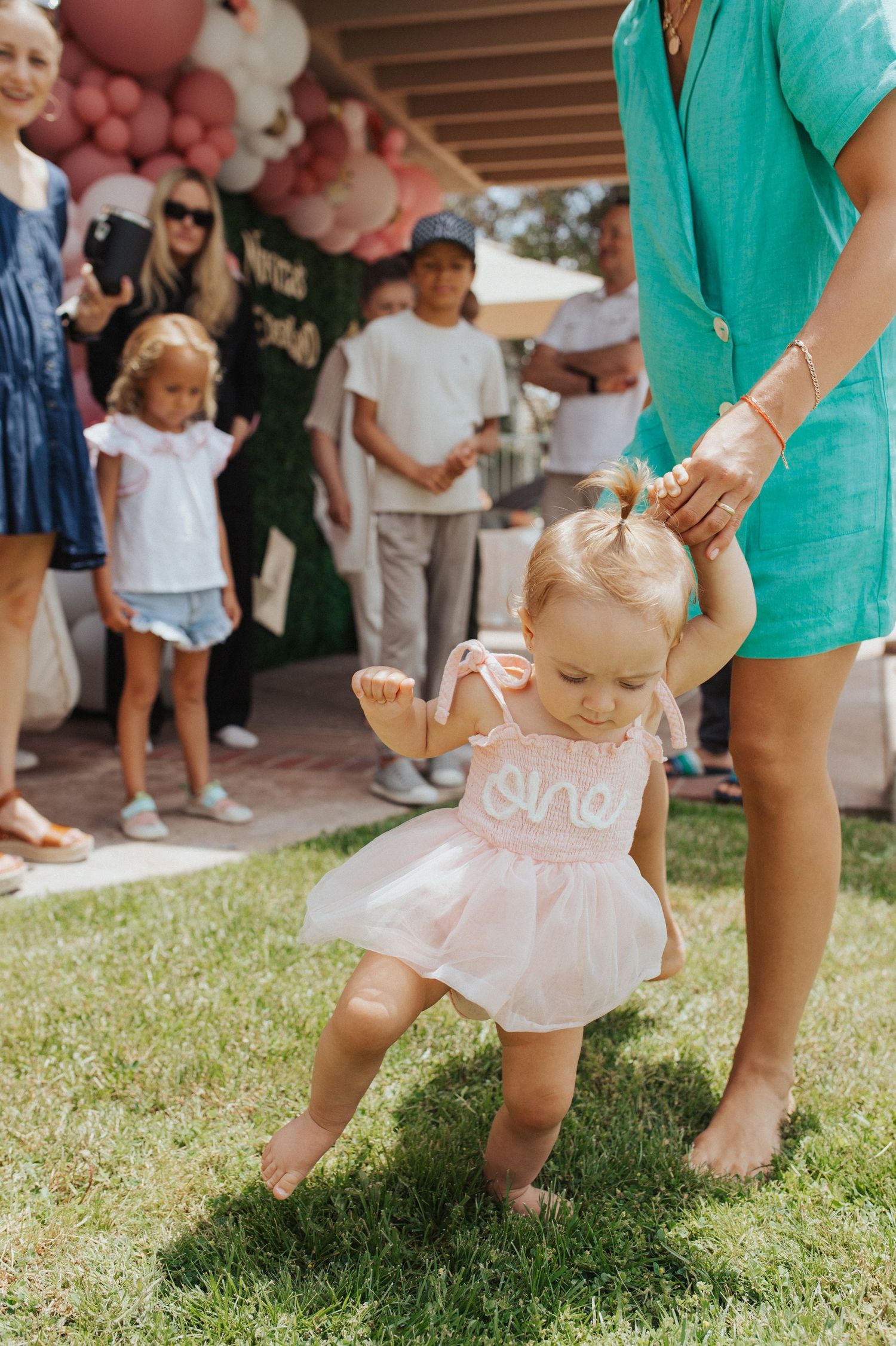 Baby girl in pink tutu dress walks on grass at party.