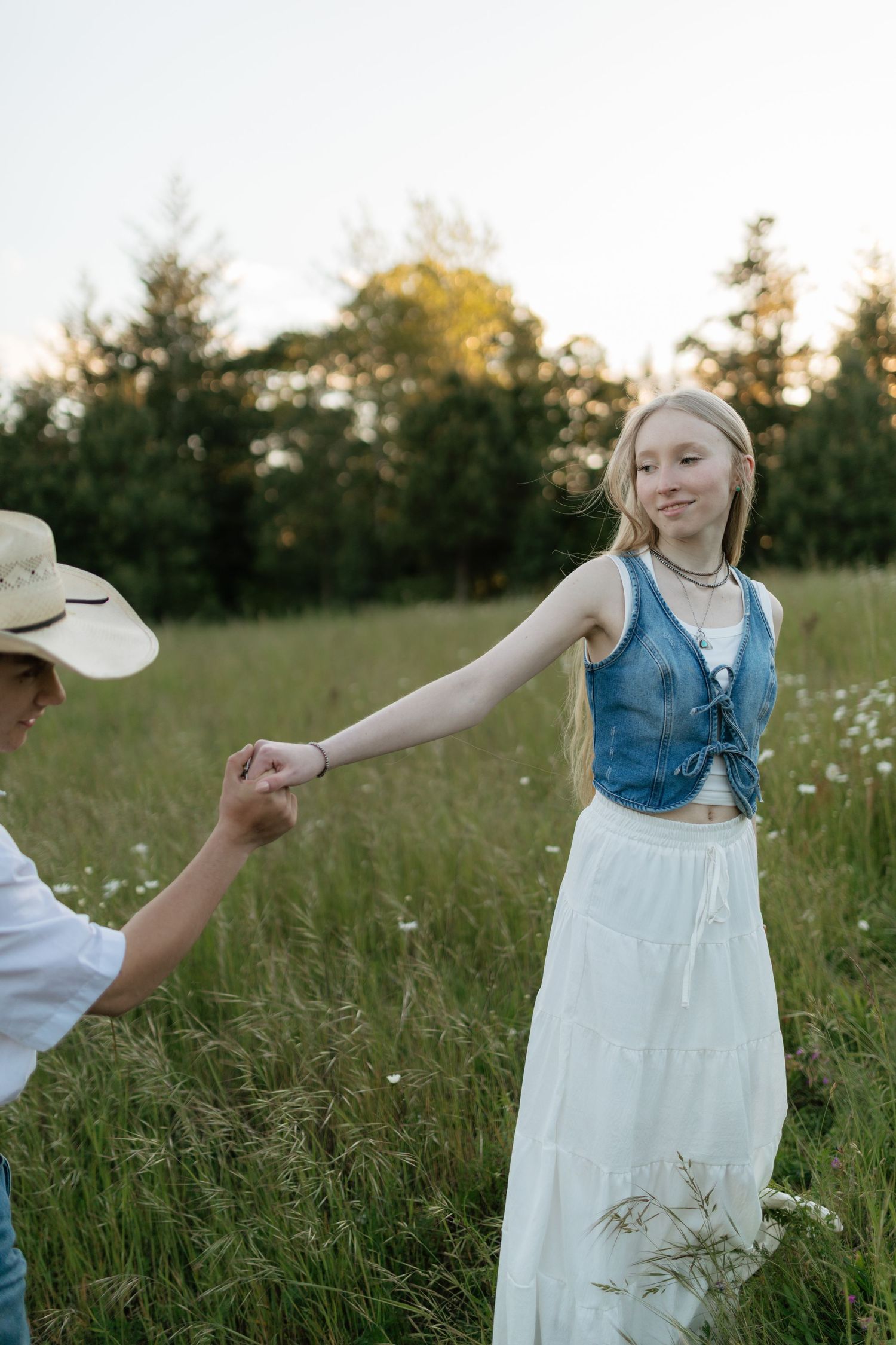 Young woman in white skirt and denim top holding hands with partner in a wildflower meadow.