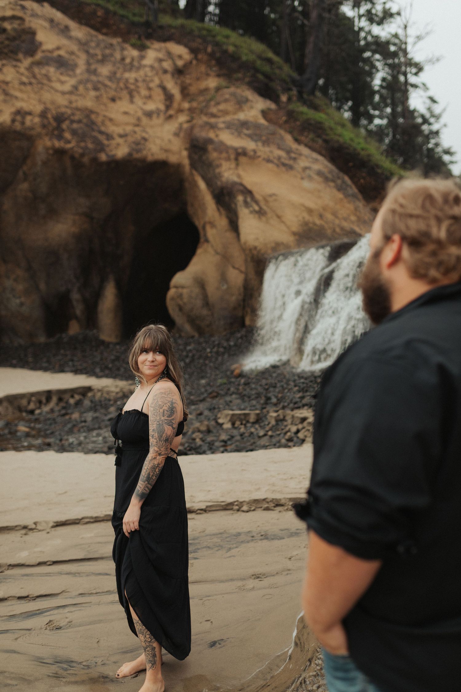 Tattooed woman in black dress poses near a beach waterfall while her bearded partner stands nearby.