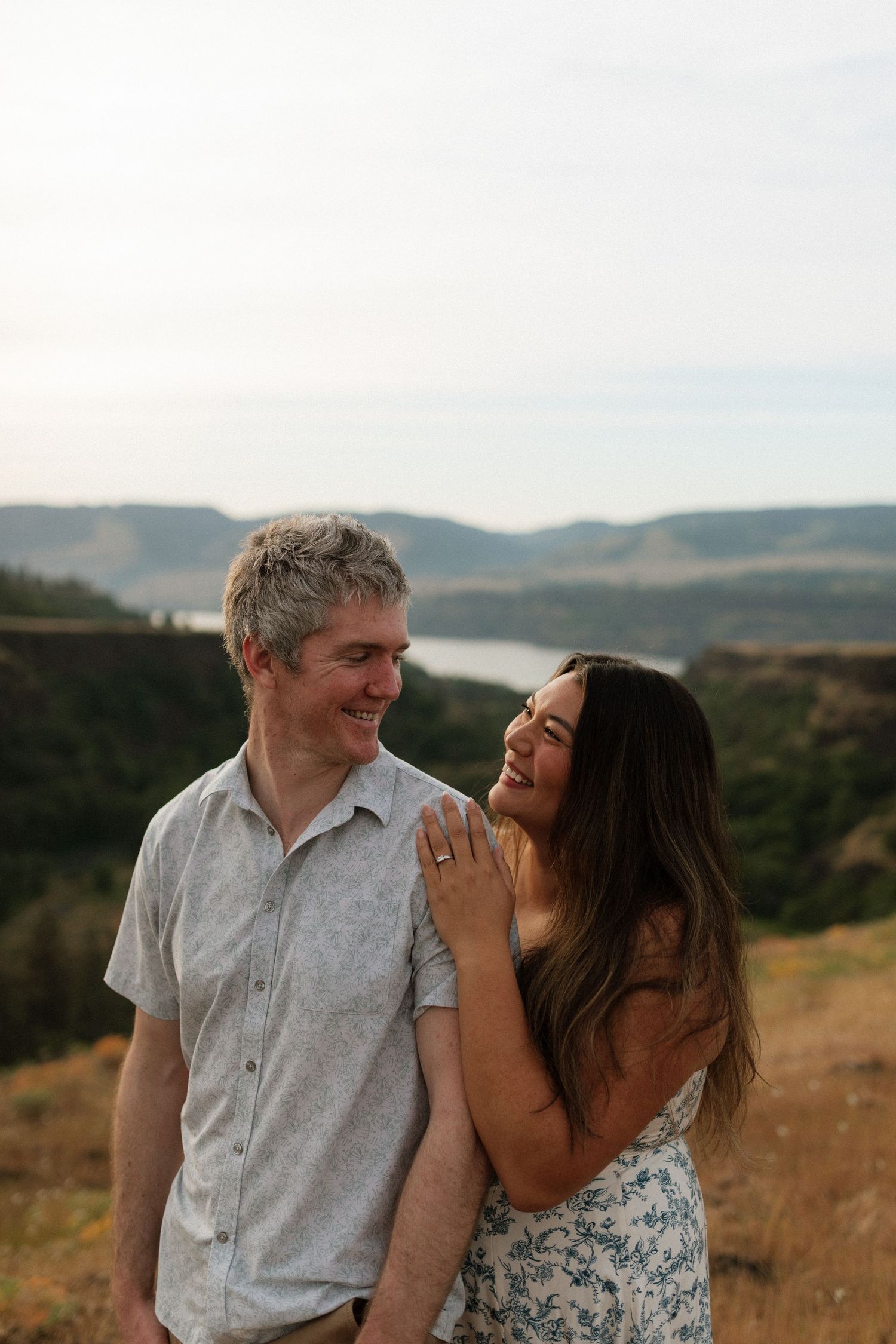 Smiling couple laughs together on hilltop with river valley and mountains in the background.