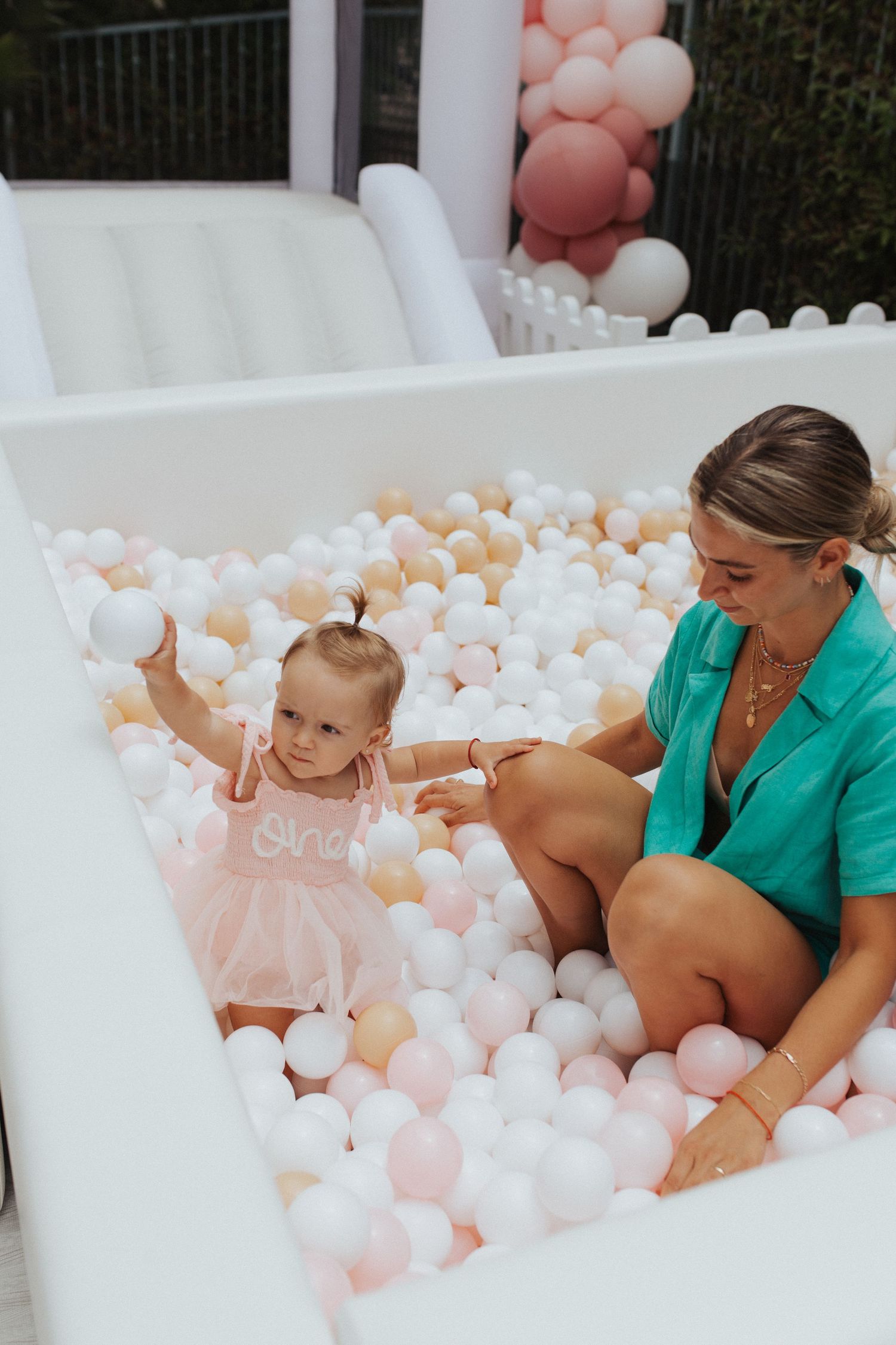 Mother in teal dress plays with toddler in pink in ball pit.