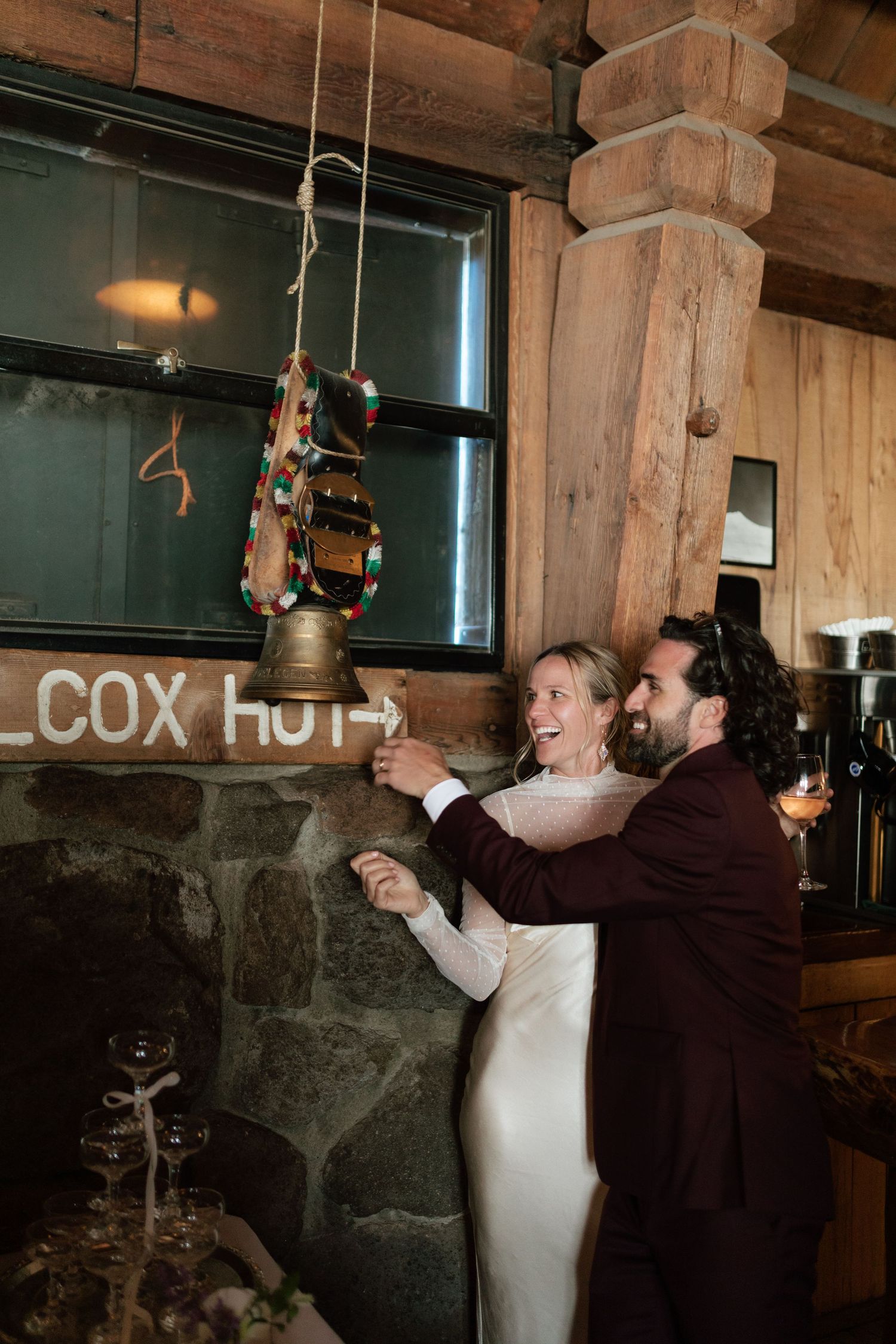 Newlyweds laugh together while ringing a decorative bell inside a cozy rustic lodge reception.