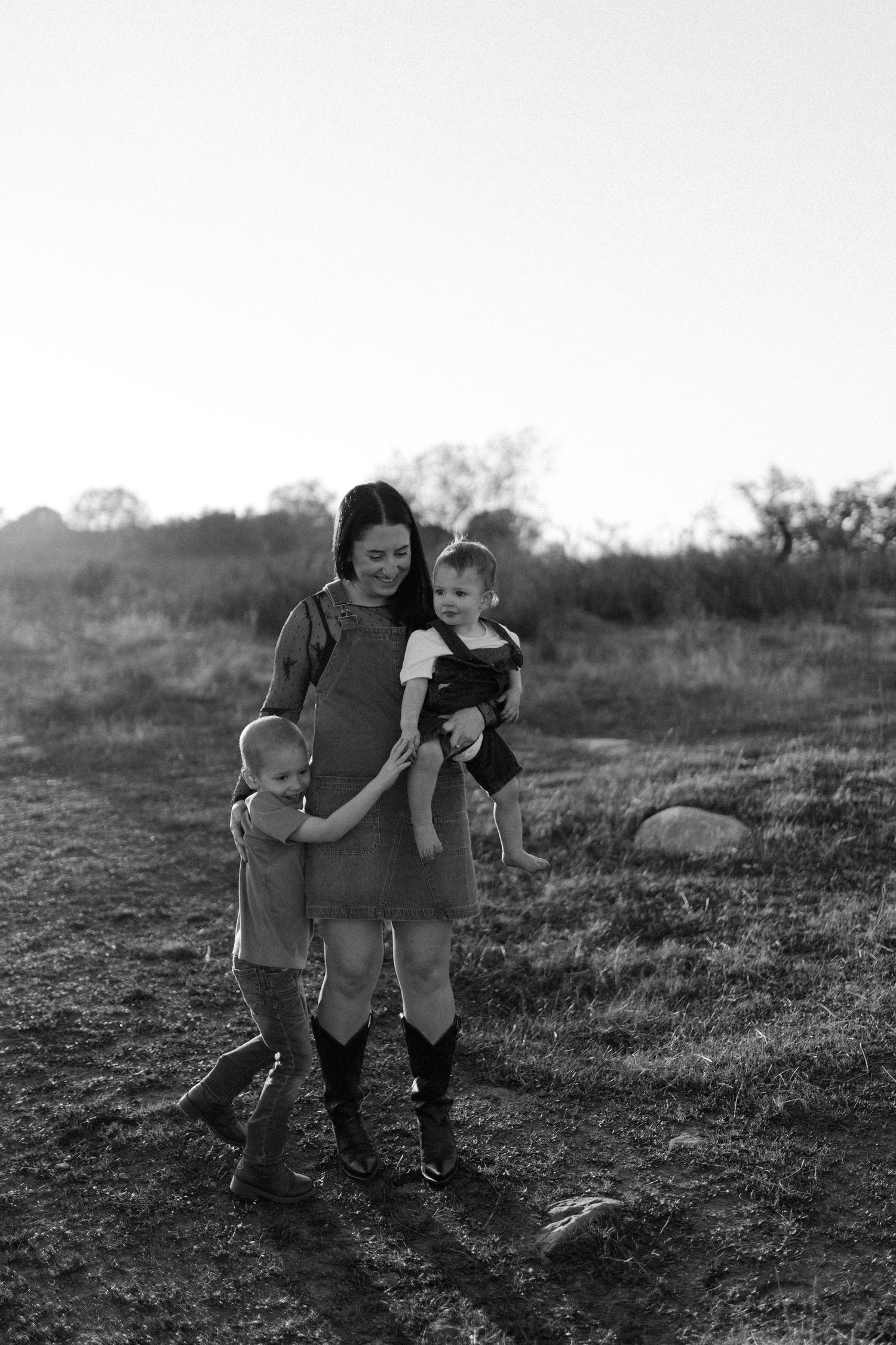 Black and white photo of mother walking with two young children through an open field at dusk.