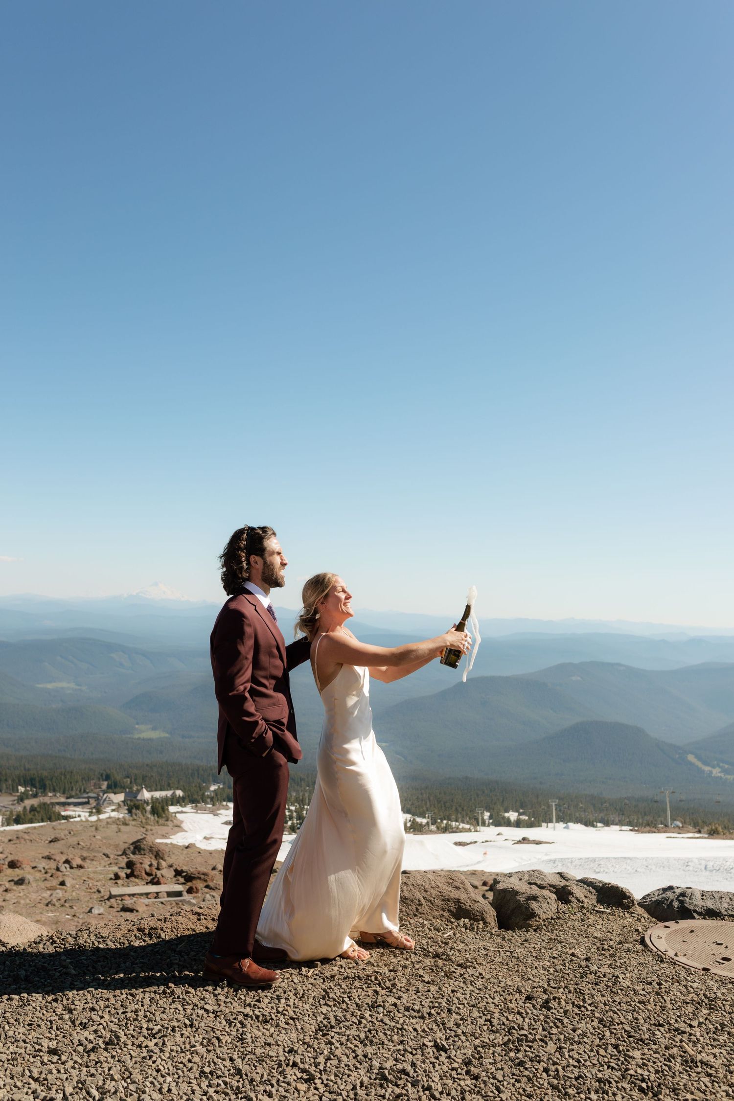 Bride pops champagne bottle with groom on scenic mountain summit on wedding day.