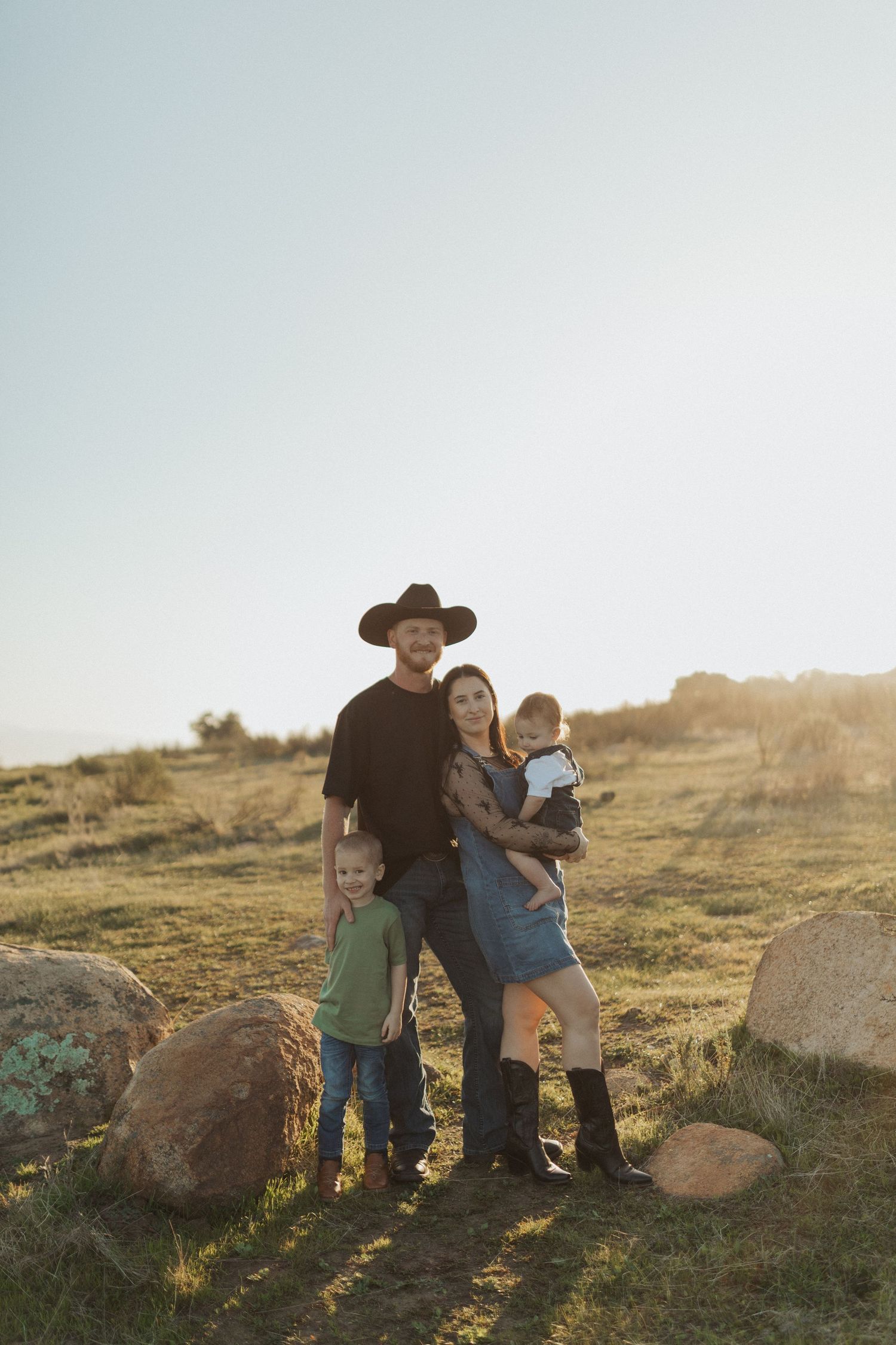 Family of four posing in sunny field, father in black cowboy hat, mother holding infant with young son.