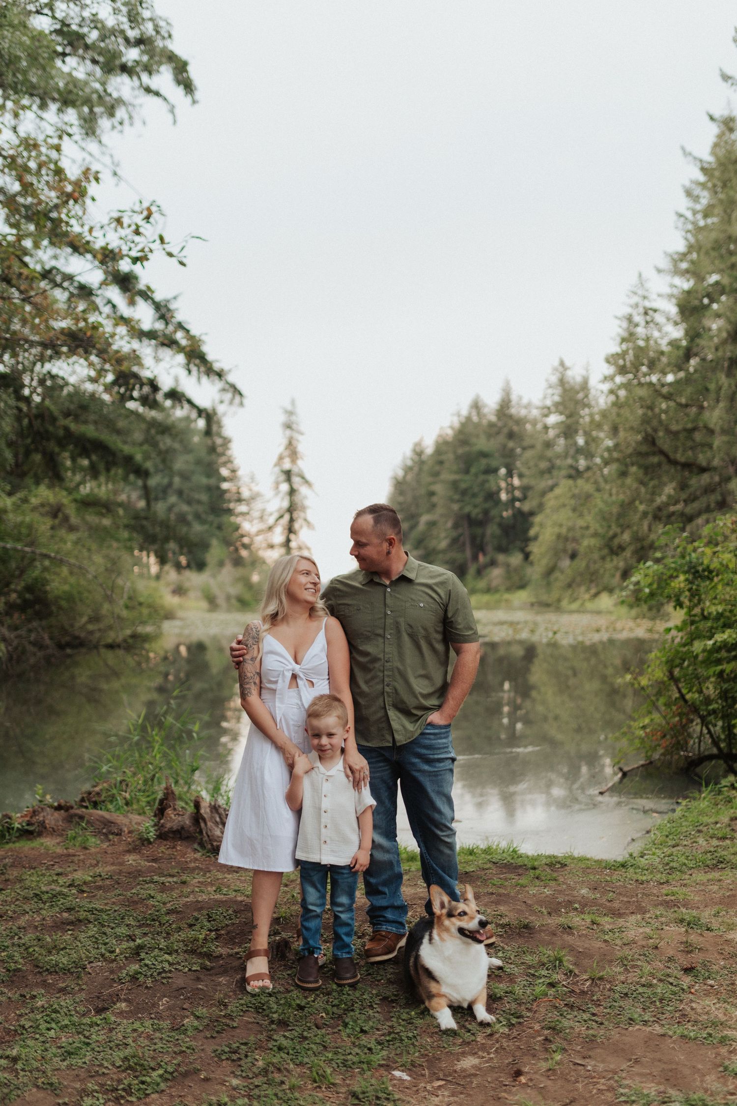 Happy family with toddler and corgi dog standing together near a serene forest river.
