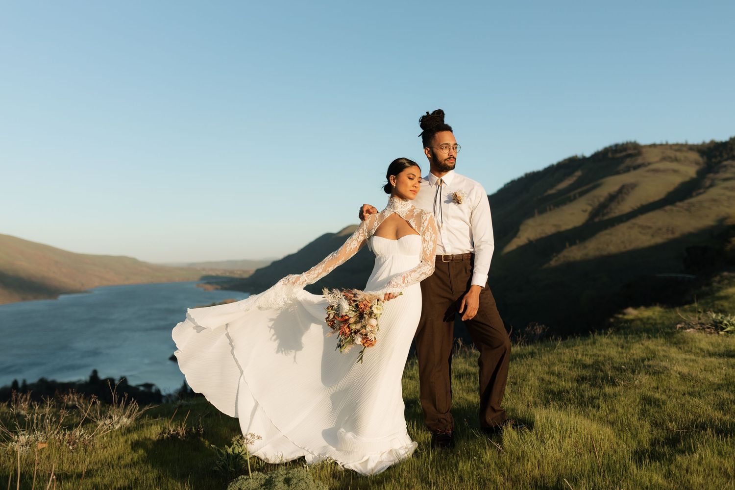 Bride in flowing white gown and groom standing on a scenic hilltop overlooking a lake and mountain landscape. Columbia Gorge Oregon Wedding.