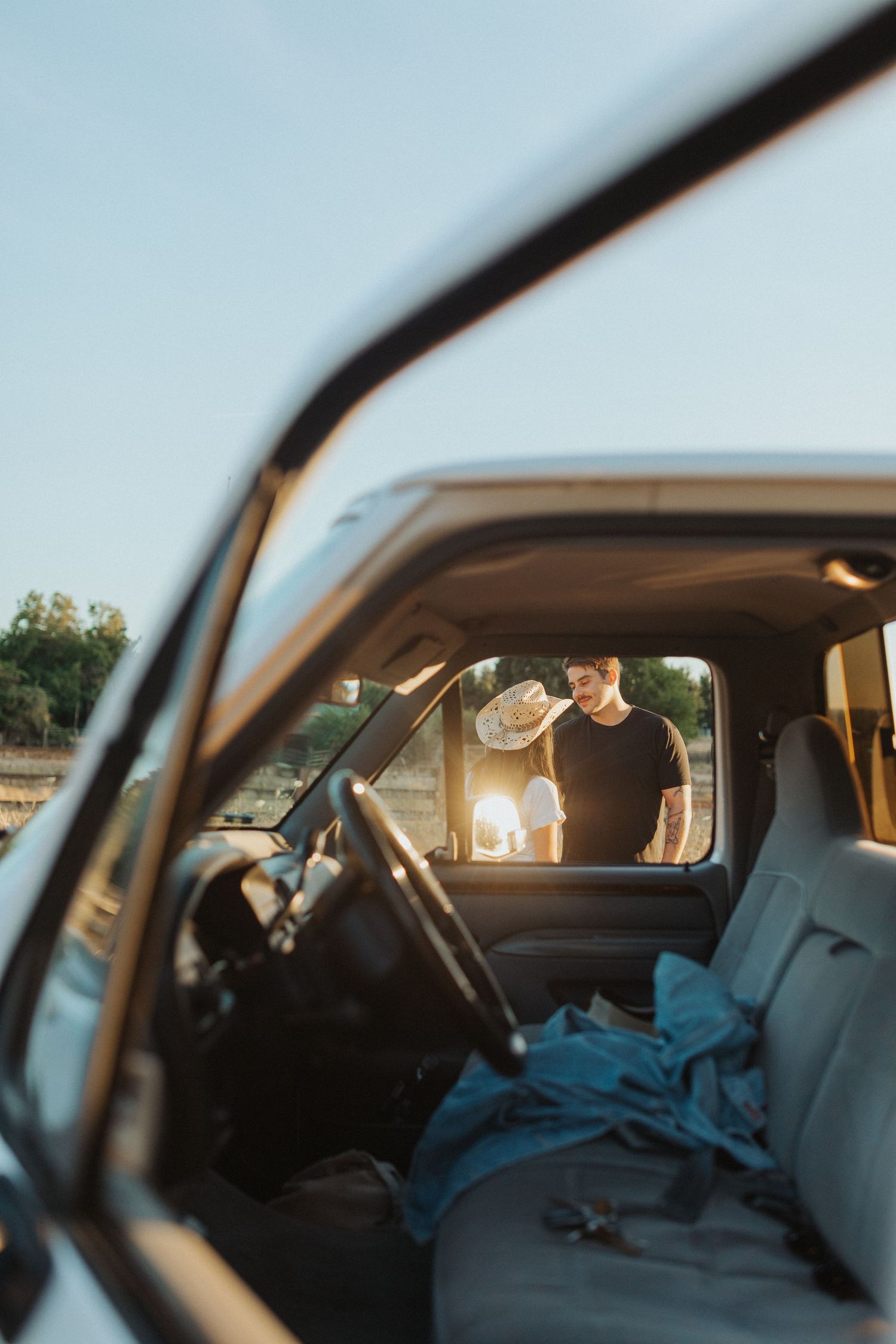 A couple stands outside a vintage truck in golden sunset light, viewed through the trucks interior window.