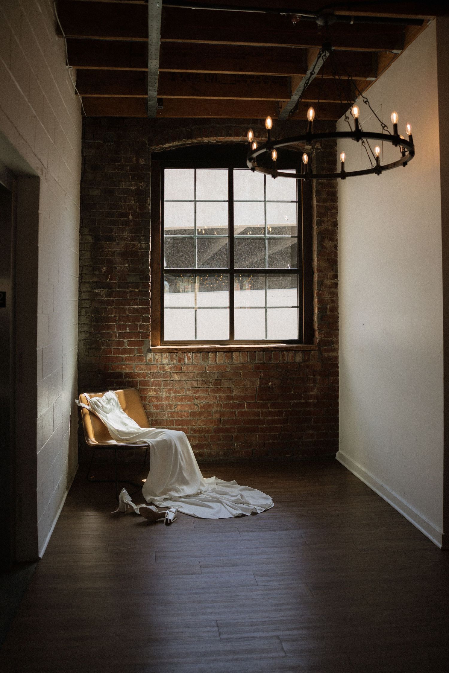 Wedding dress draped over a chair in a rustic brick-walled venue with chandelier lighting.