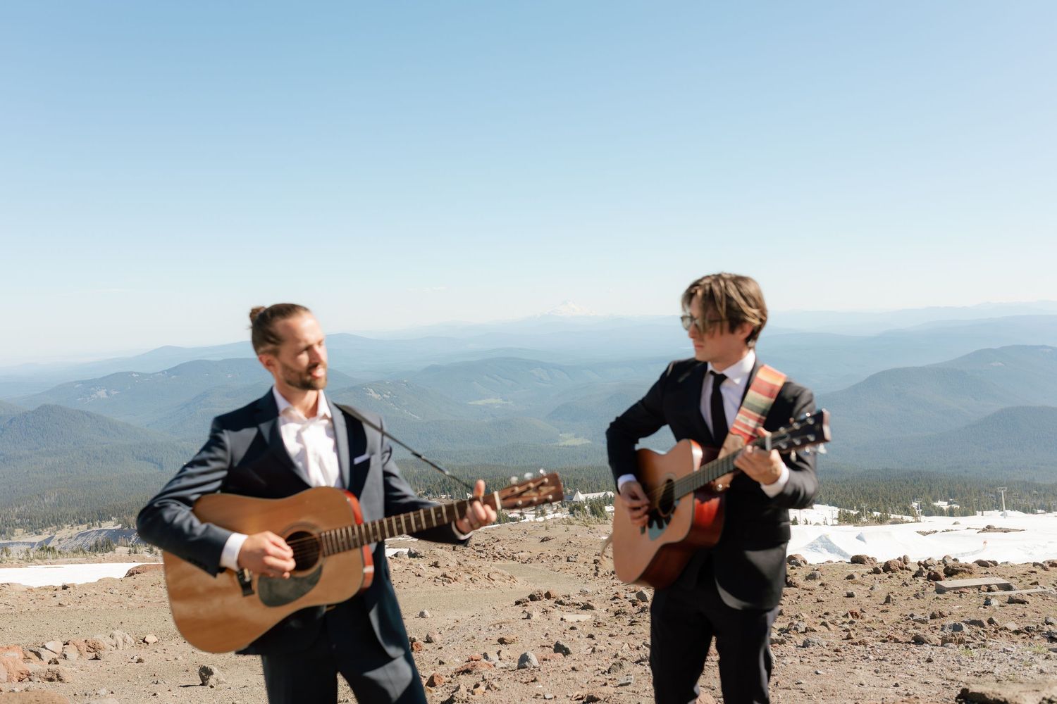 Two musicians playing acoustic guitars on rocky mountaintop with panoramic views.