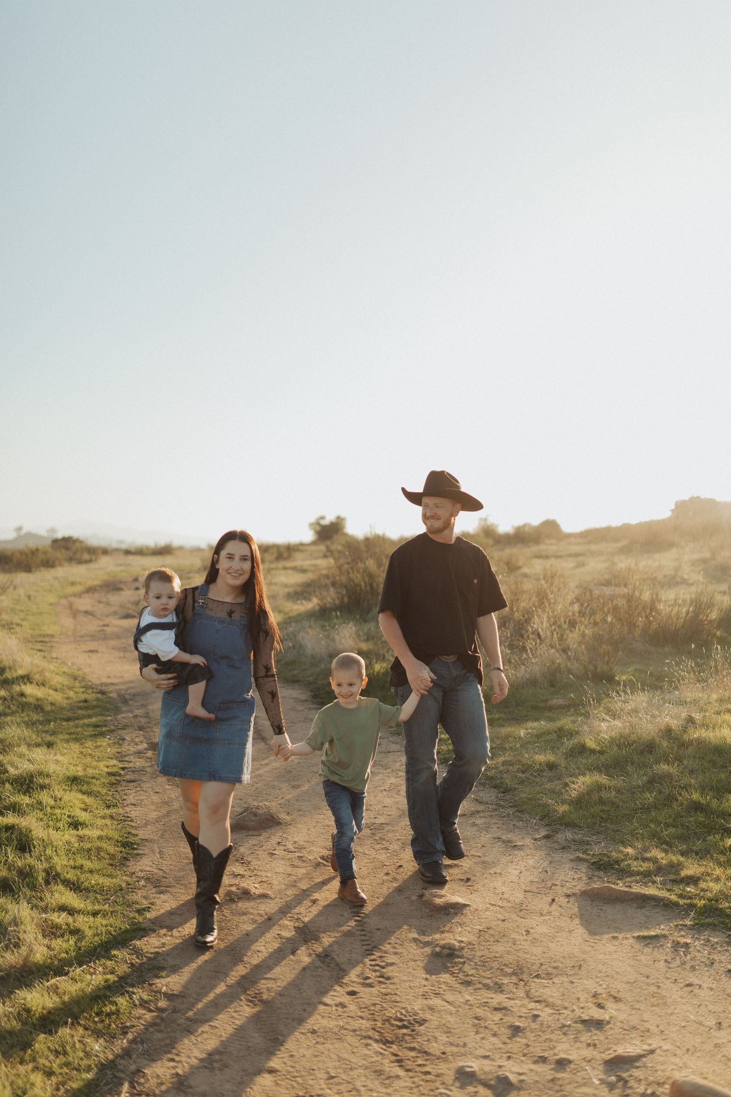 Family of four walking dirt path in sunny field, mother holding baby, father in black cowboy hat.