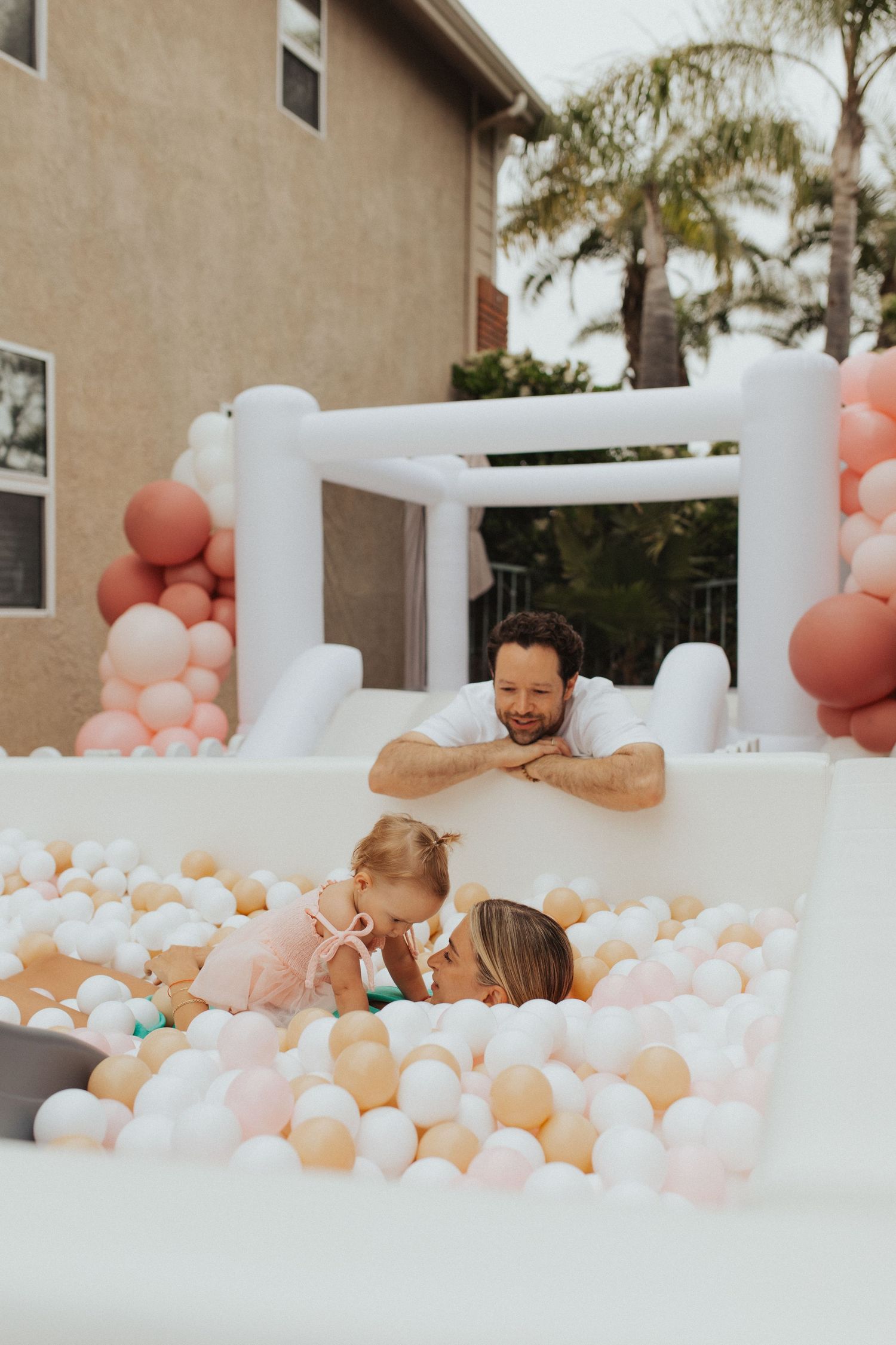 Father watches mother and baby play in outdoor ball pit.