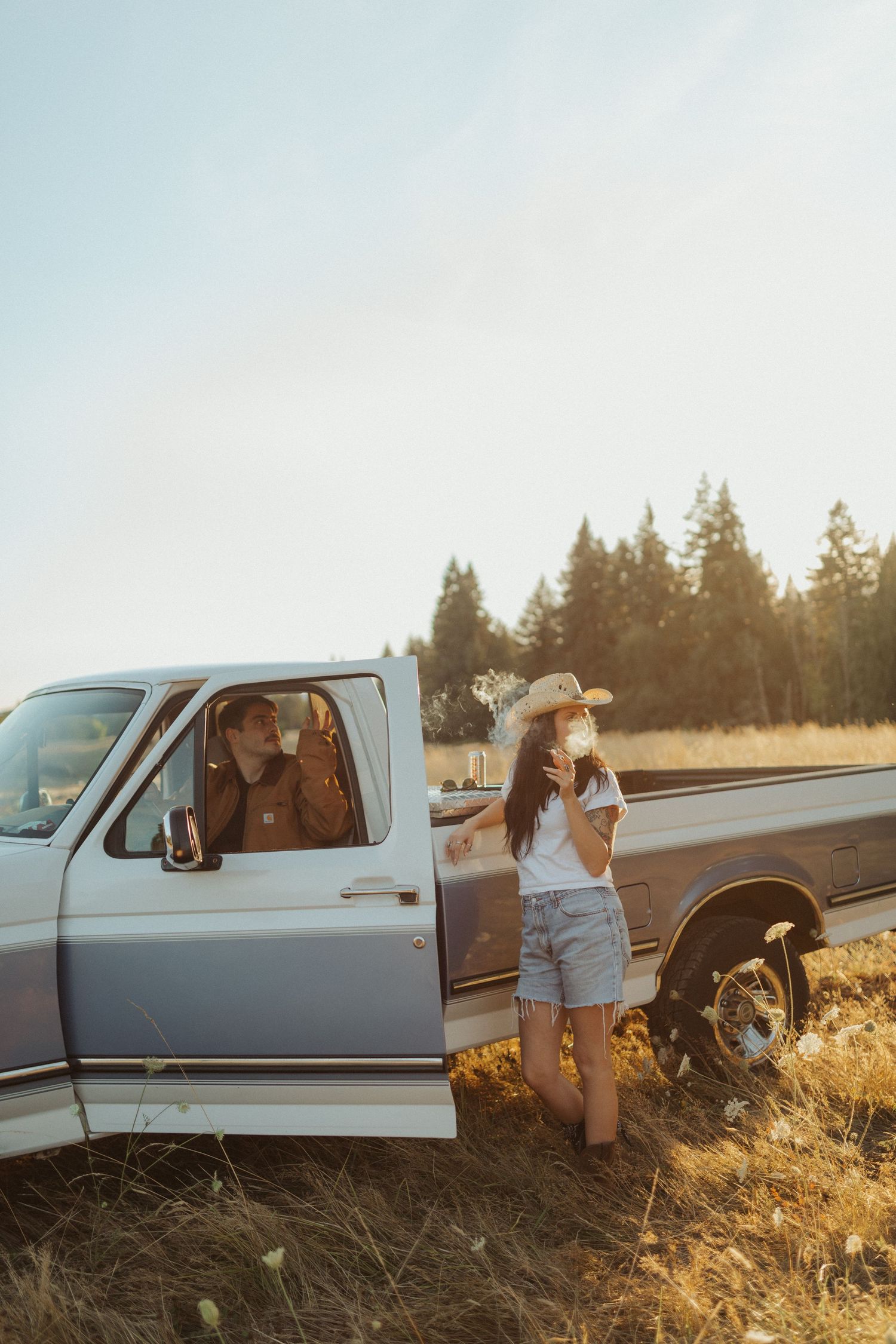 A woman in a cowboy hat holds wildflowers beside a vintage blue pickup truck in a golden field at sunset.