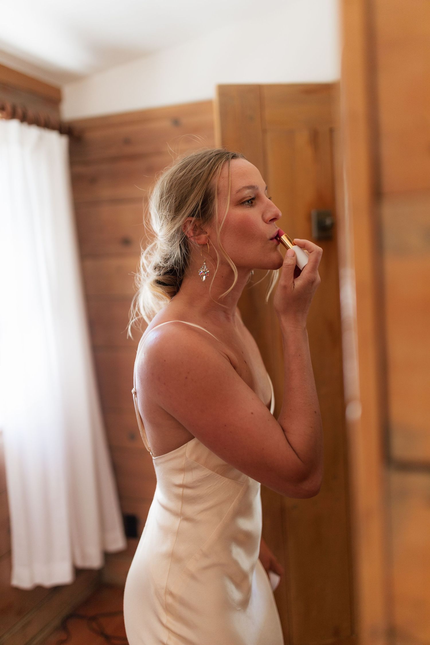 Blonde bride in elegant satin slip wedding dress applies lipstick while getting ready in a wood-paneled room.