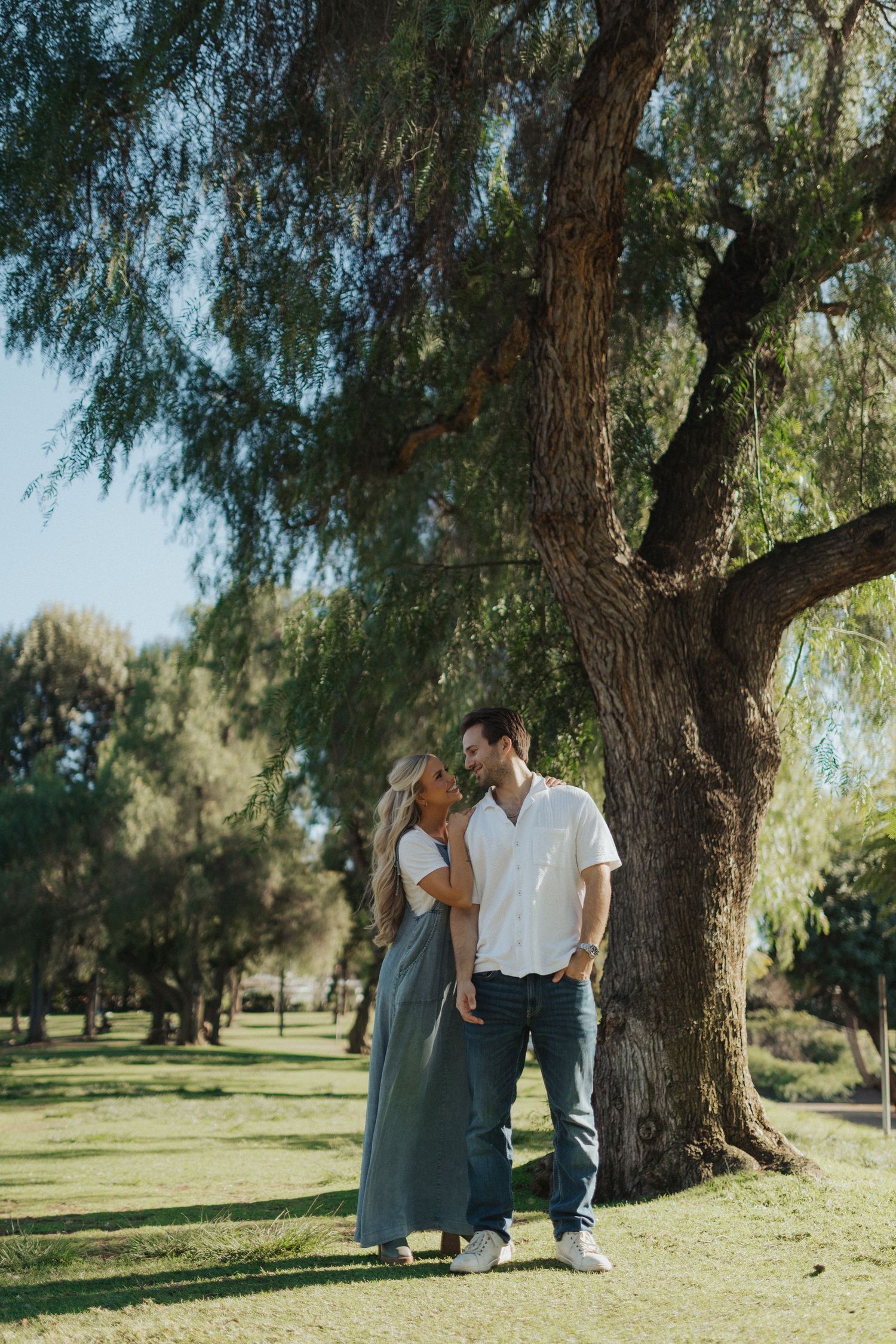 A romantic couple shares a tender moment standing beneath a large tree in a sunny green park.