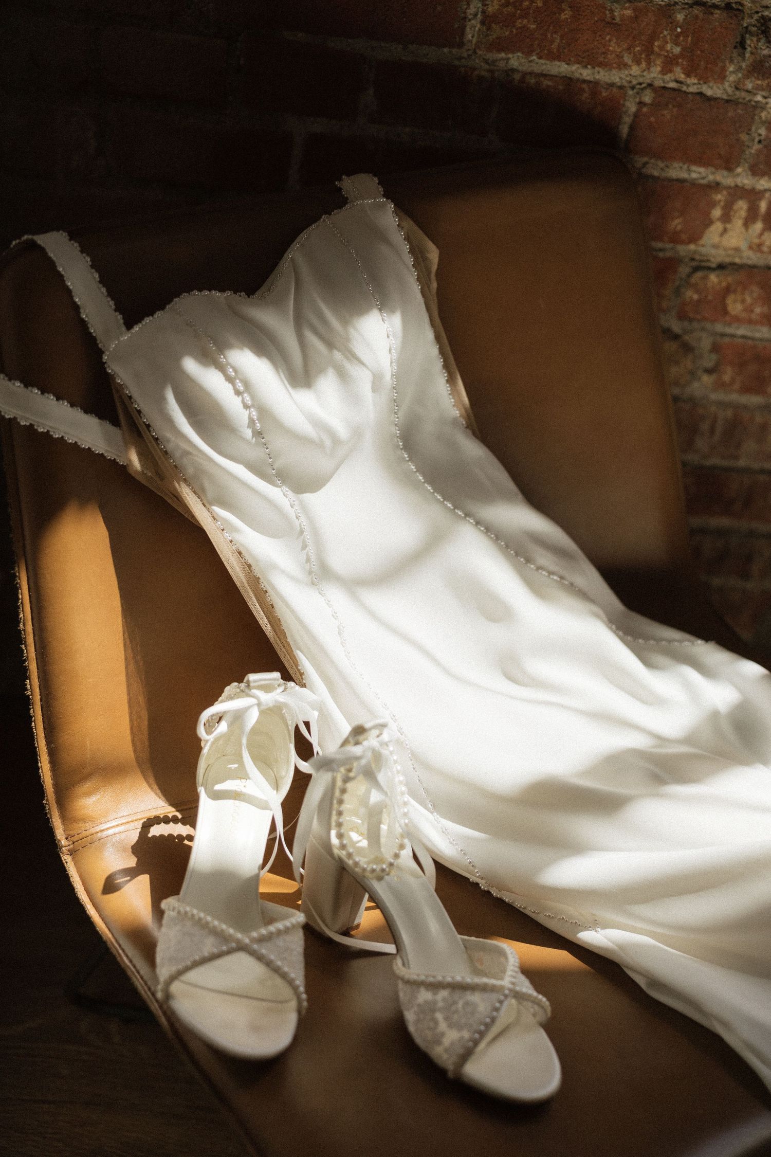 White wedding dress and pearl-embellished heels displayed on a vintage chair against a rustic brick wall.