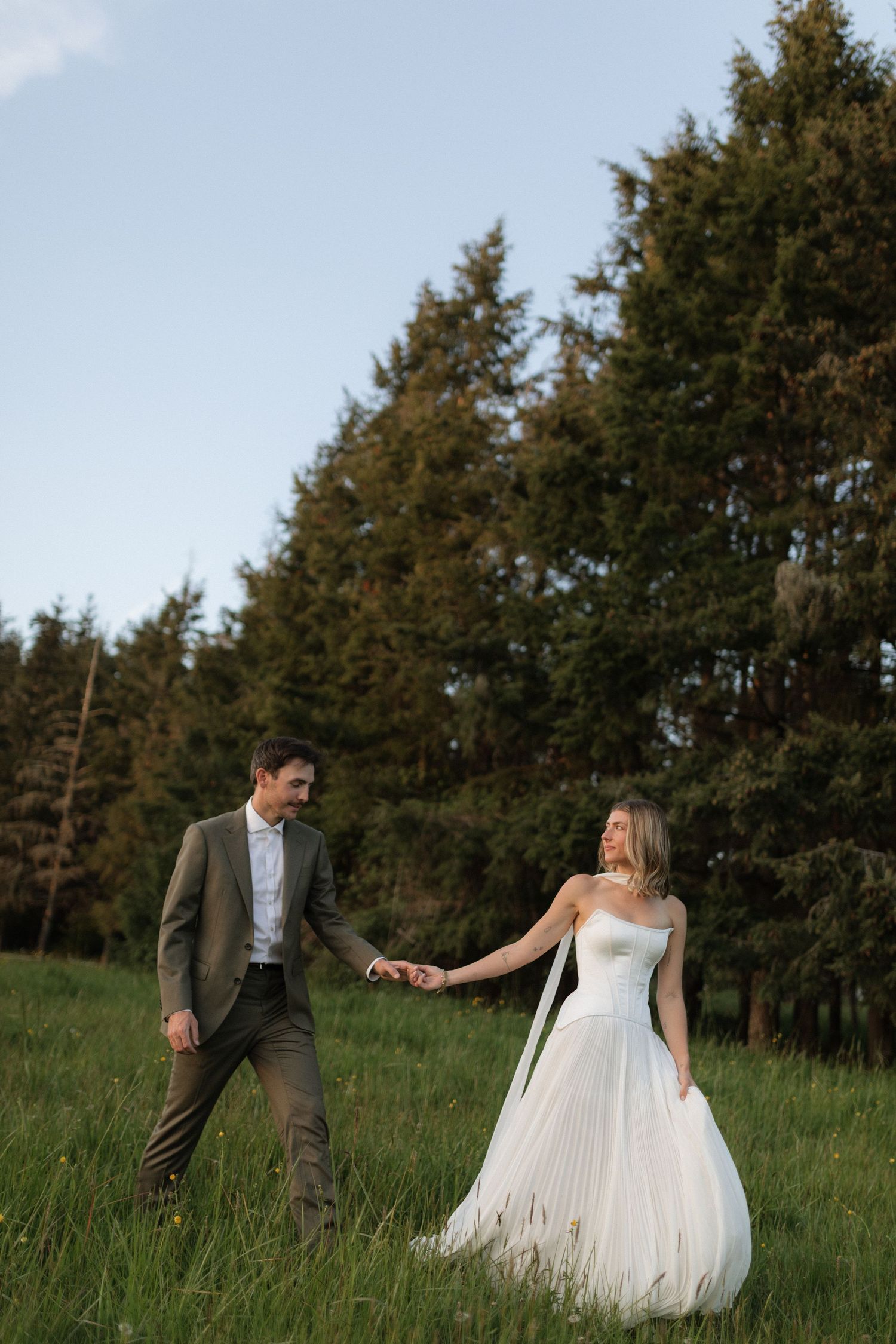 Bride in white tulle gown holding grooms hand while walking through green meadow near pine trees.
