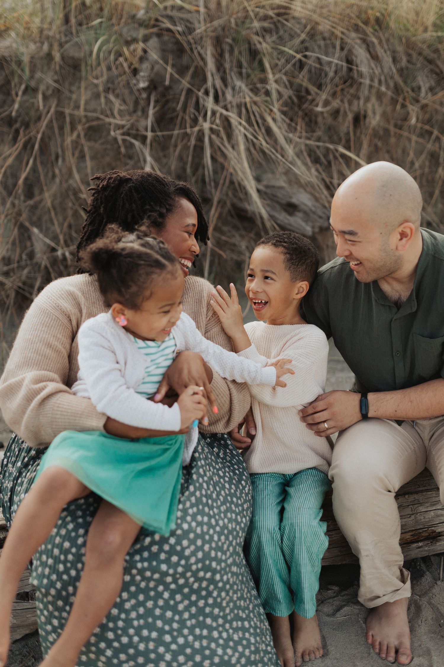 Happy interracial family of four sitting on driftwood at the beach, parents laughing with two young children.