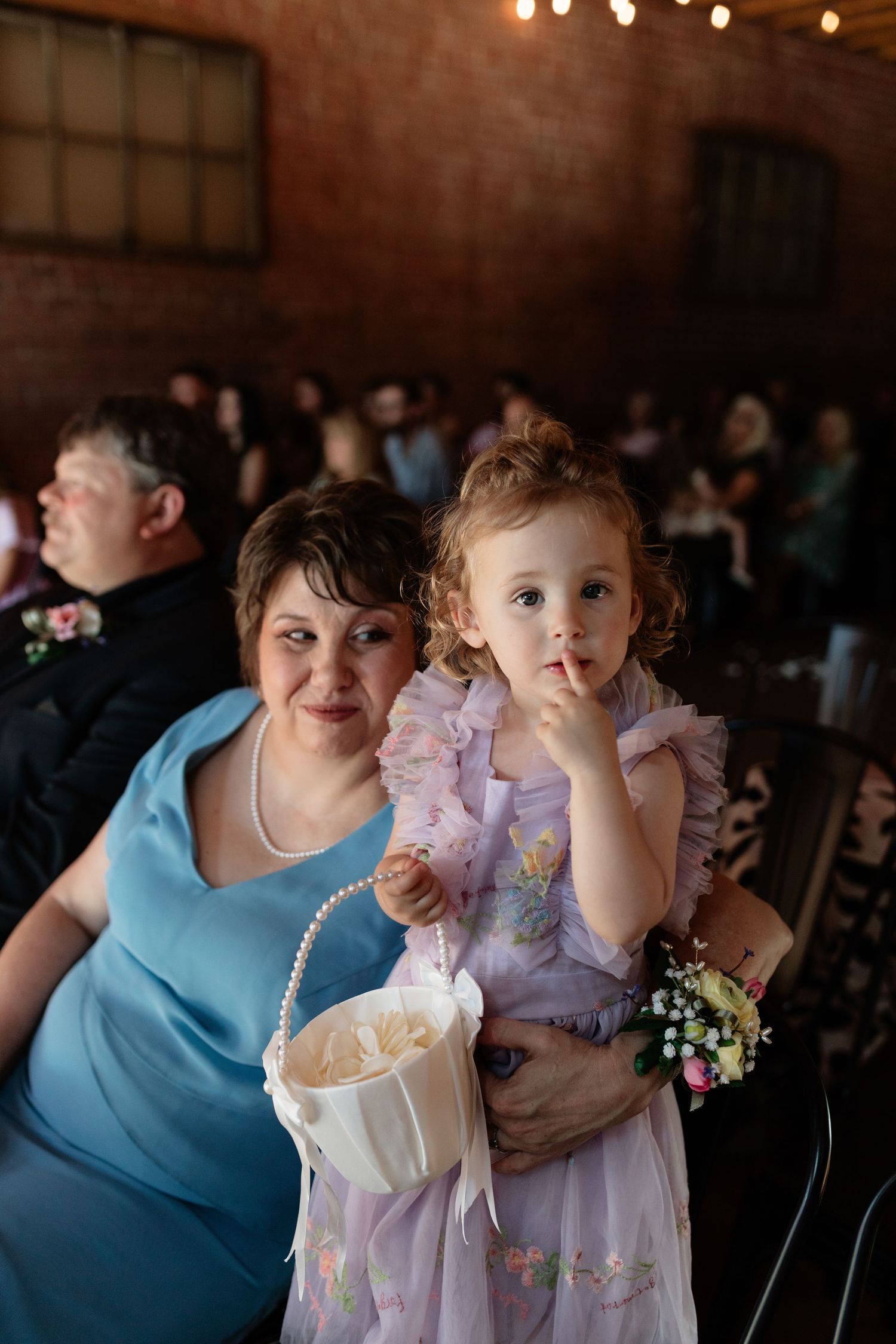 Smiling woman in blue dress holds young flower girl in purple dress during wedding ceremony.