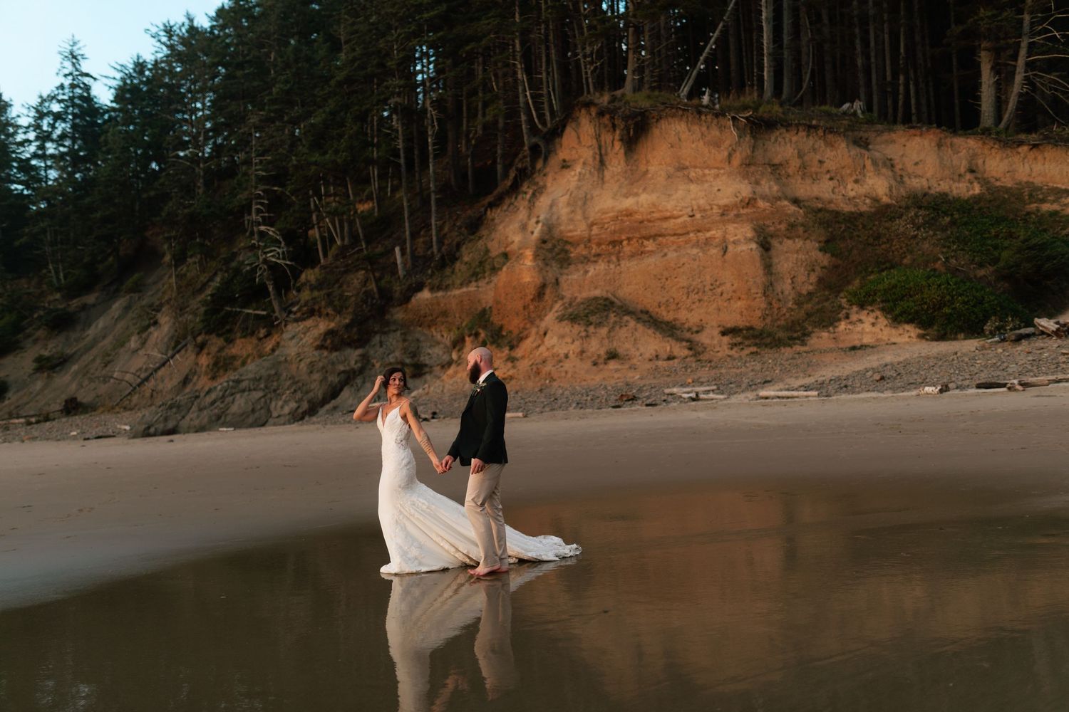 Bride and groom walk hand in hand on a reflective beach with sandy cliffs and pine trees at sunset.