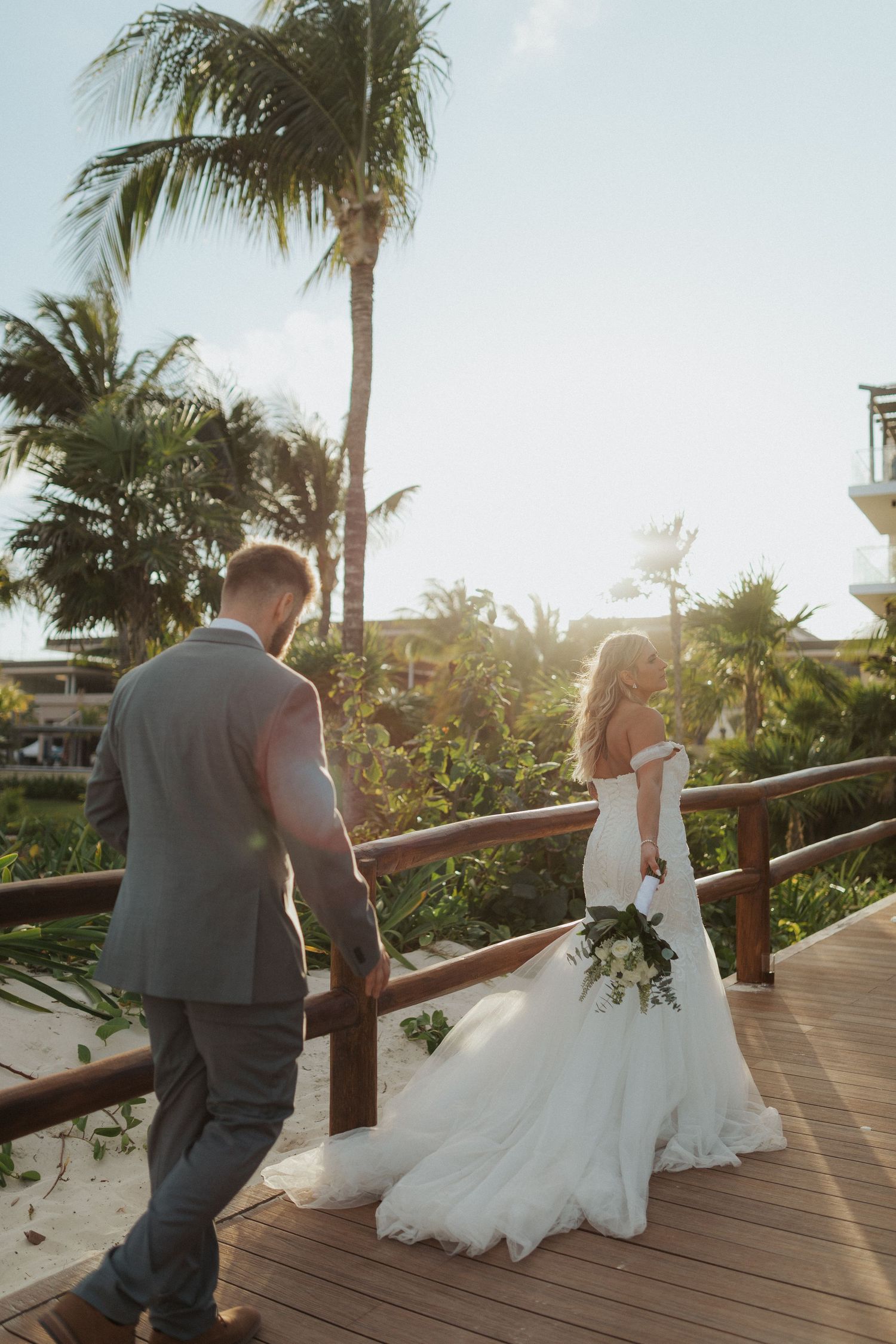 Newlyweds walking hand in hand on wooden boardwalk surrounded by tropical palms at golden sunset.