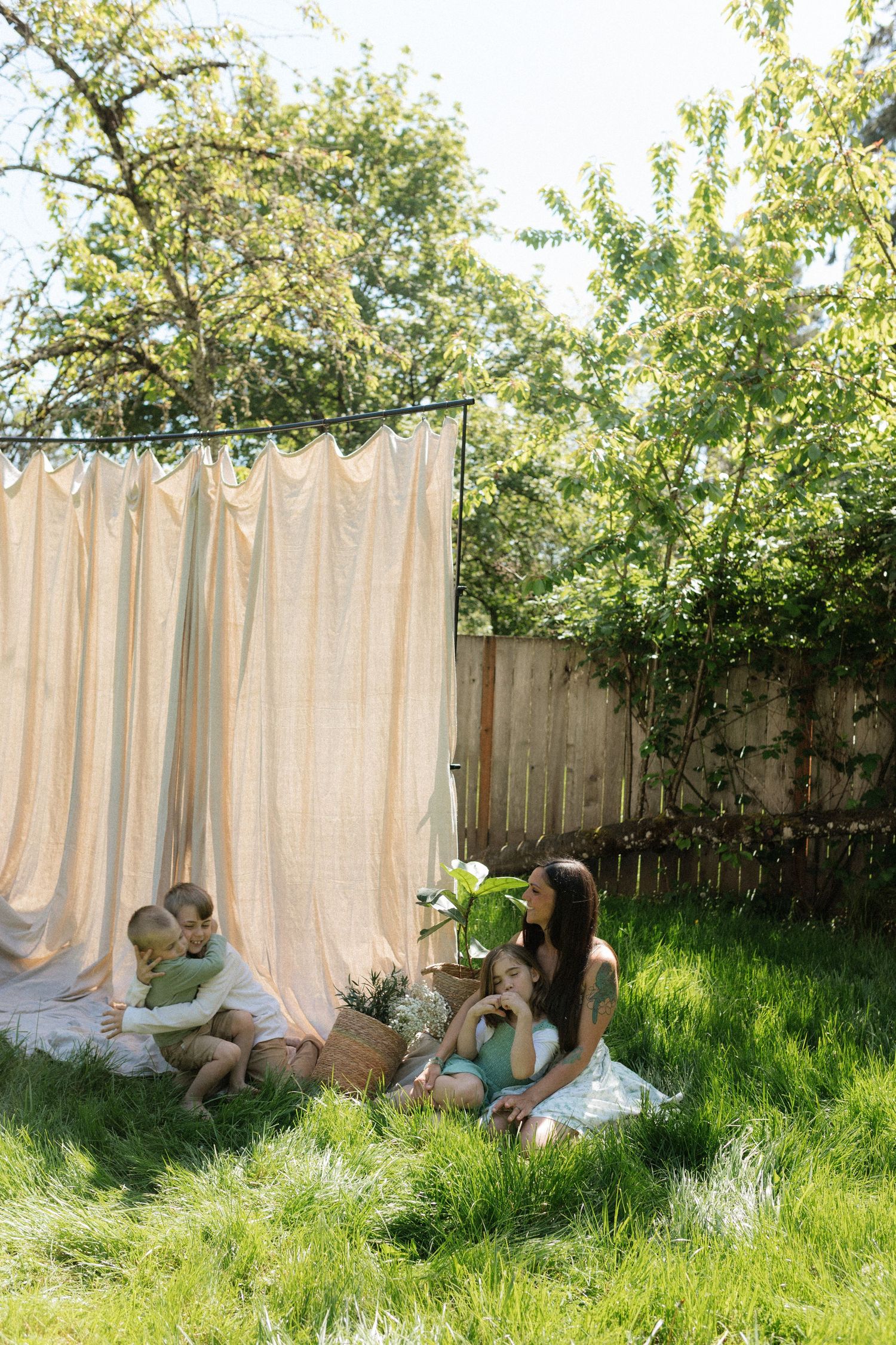 A mother and children relax on green grass beside a flowing beige curtain backdrop outdoors.