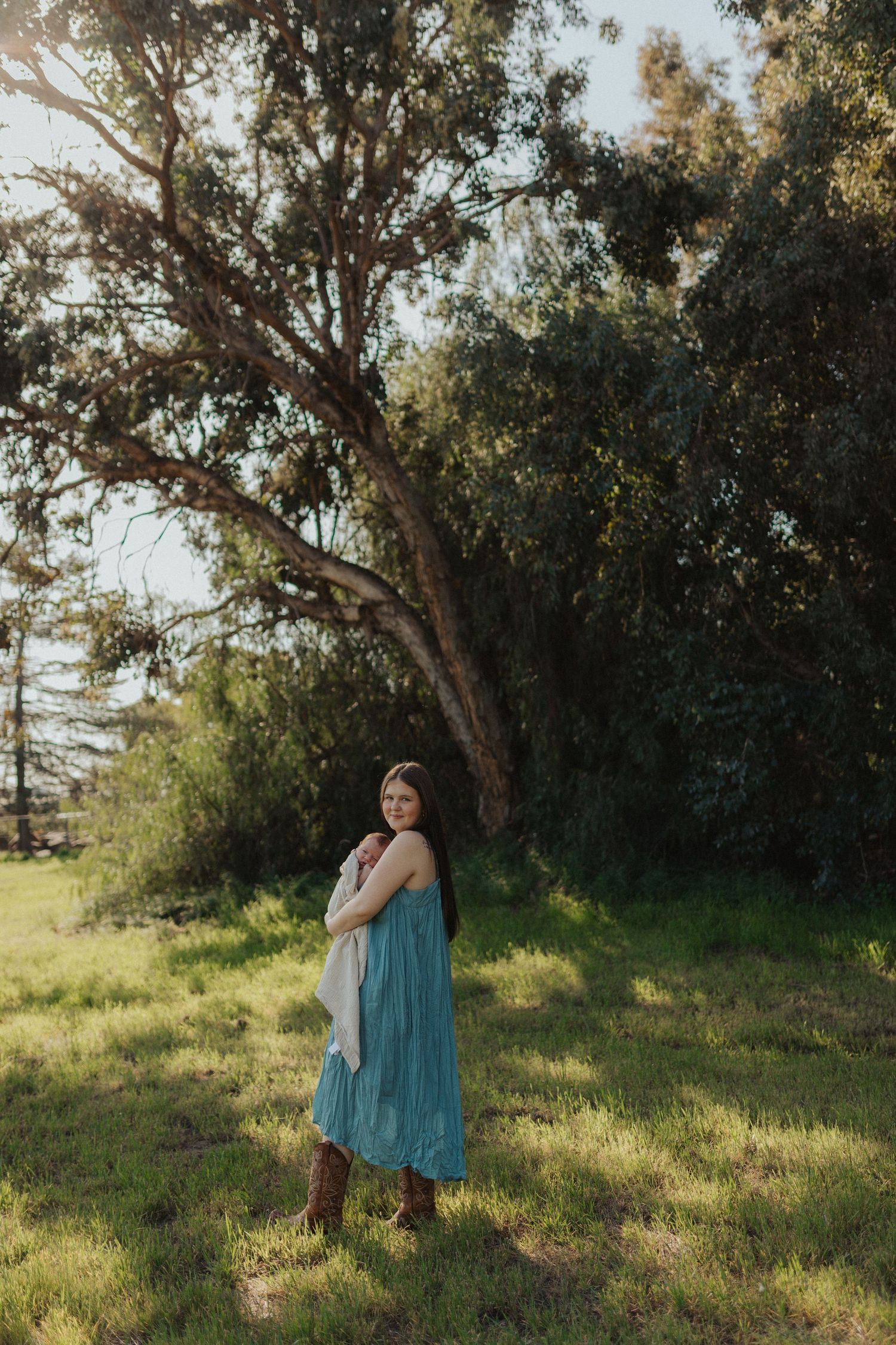 Young woman in teal dress standing in sunlit green field beside large tree holding white wrap.