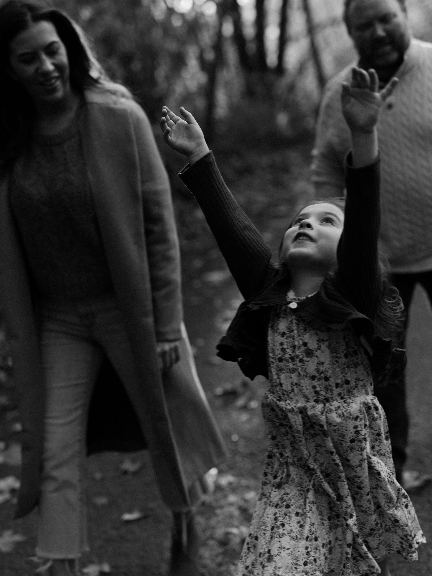 Black and white photo of a young girl reaching upward with parents smiling behind her in autumn park.