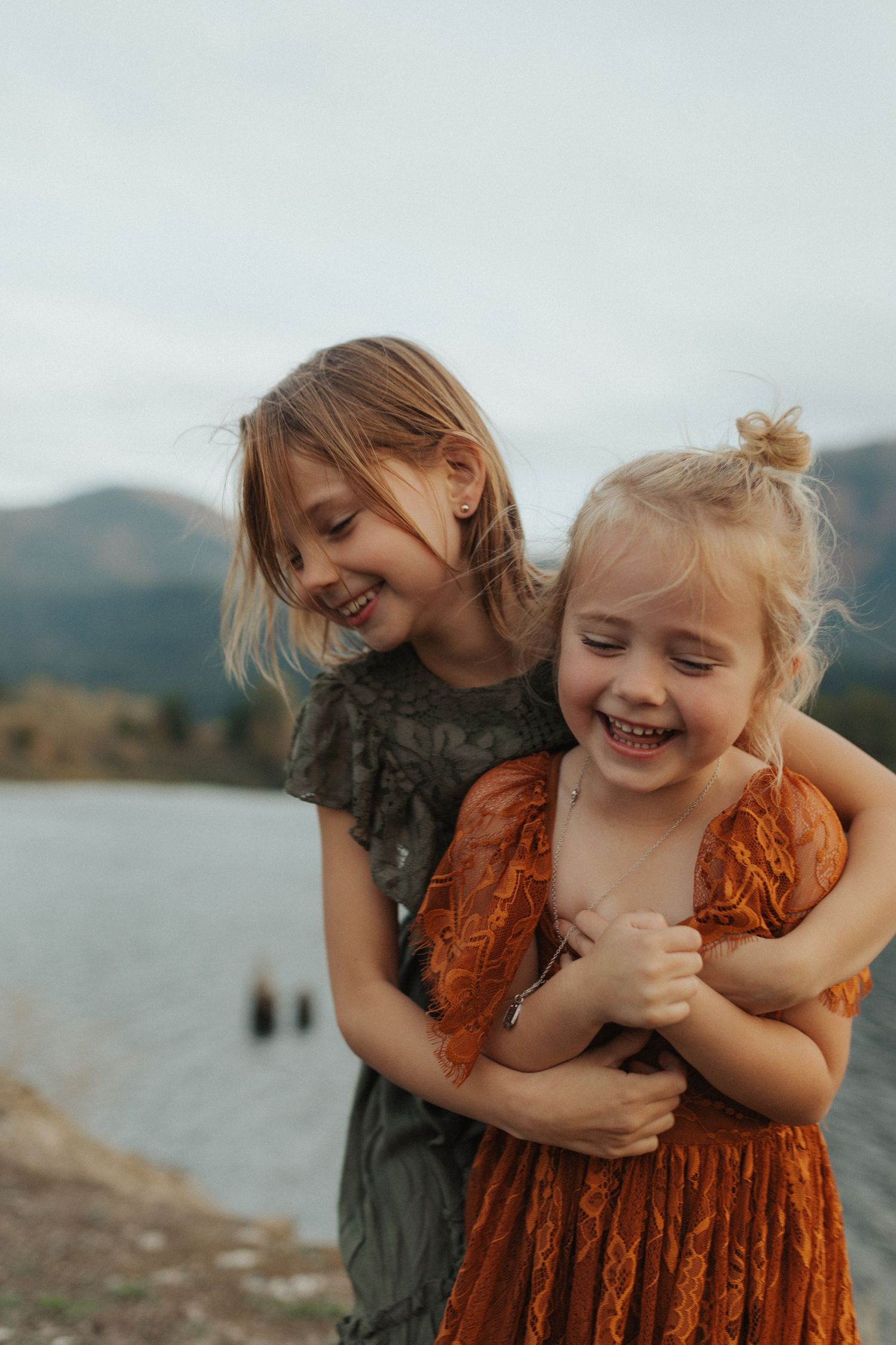 Two young sisters laugh and embrace joyfully by a mountain lake, wearing olive and rust dresses.