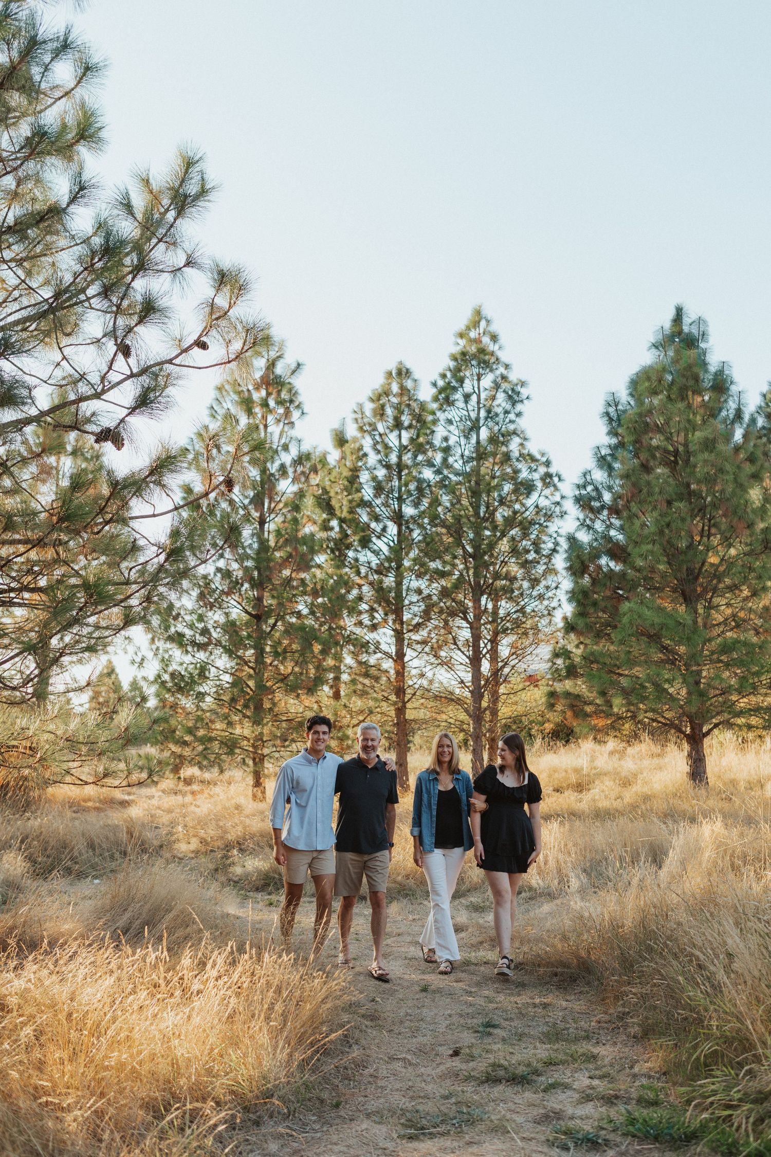 Four adults walk together on a dirt path surrounded by tall pine trees in golden field.