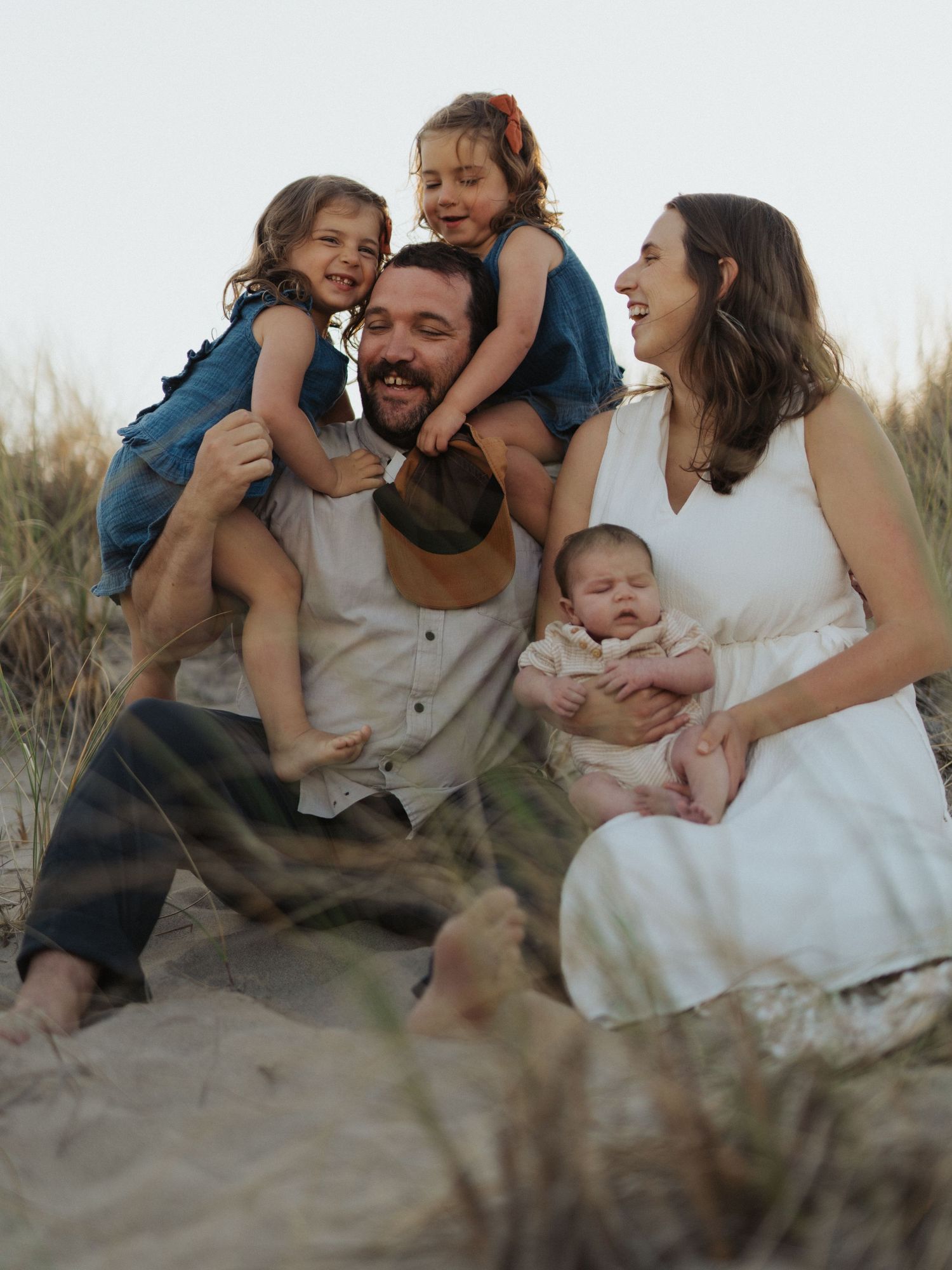 Joyful family of five with dad, mom, newborn, and two daughters laughing together on sandy beach dunes.