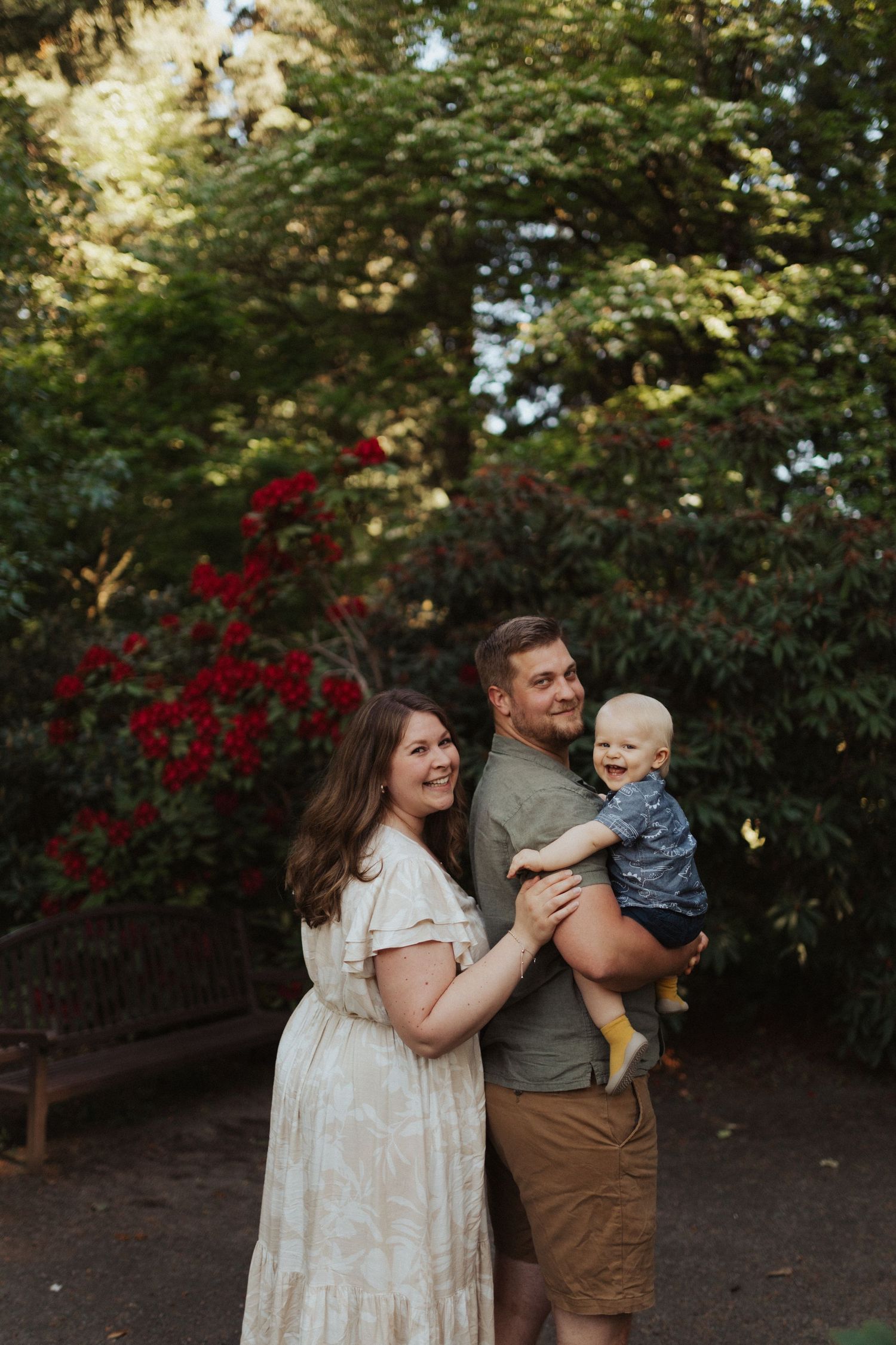 Happy family of three with laughing toddler posing together in lush green garden with red roses.