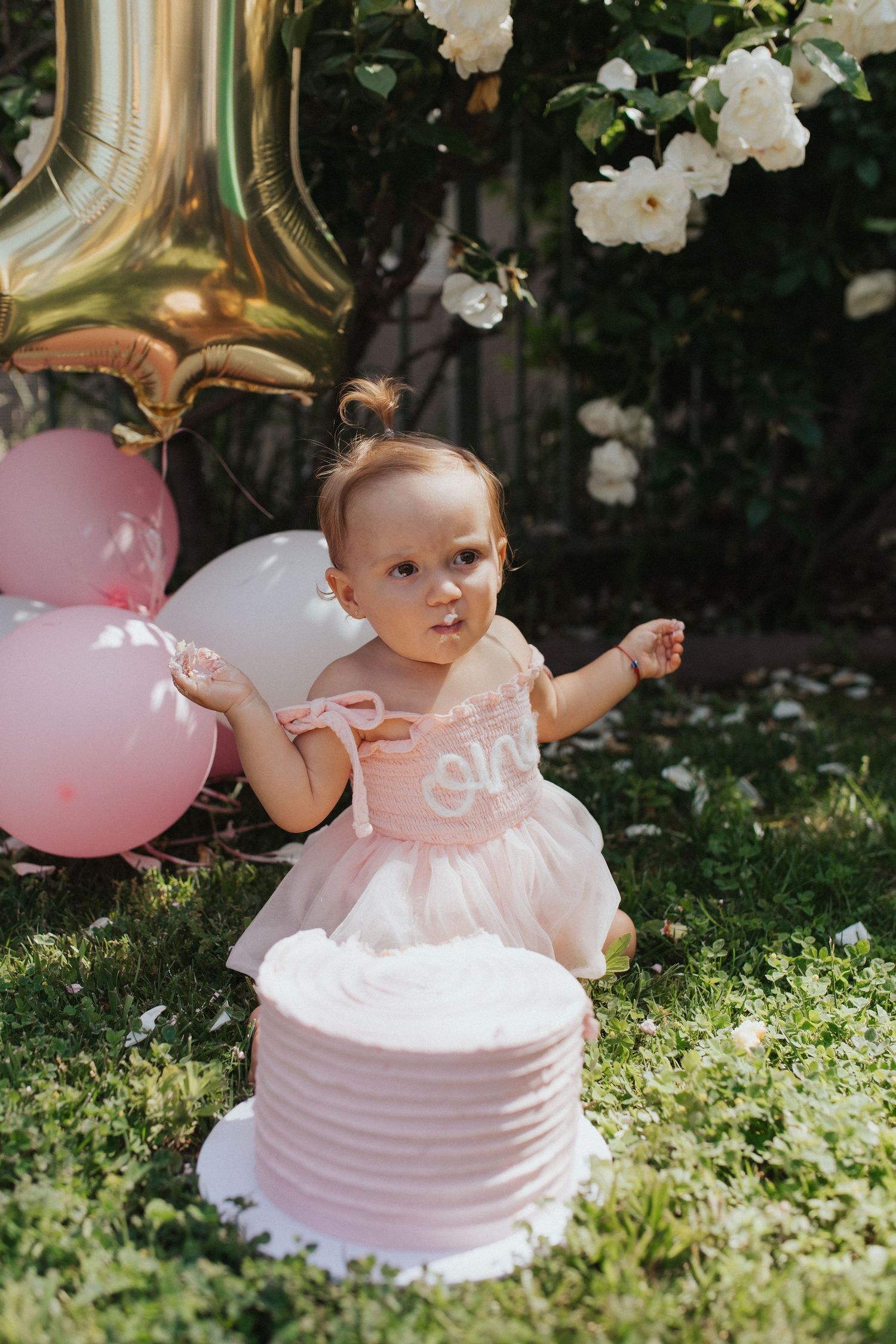 Baby in pink tutu dress reaches toward white smash cake surrounded by pink balloons outdoors.