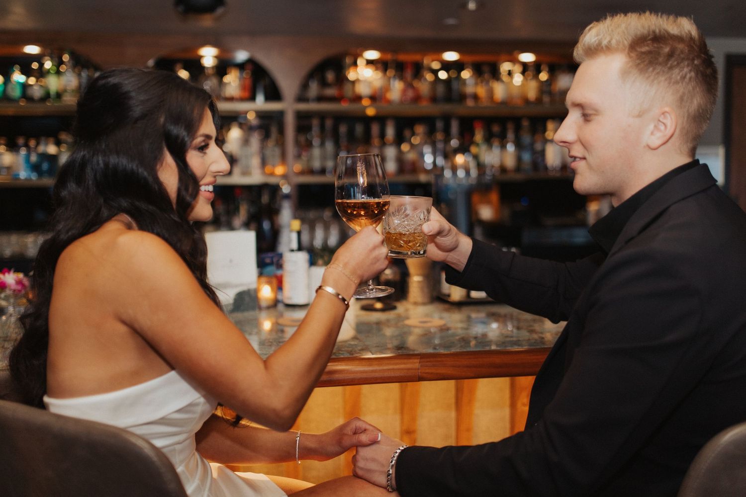 Elegant couple toasting cocktails at a luxury bar, woman in white strapless dress smiling at man in black suit.