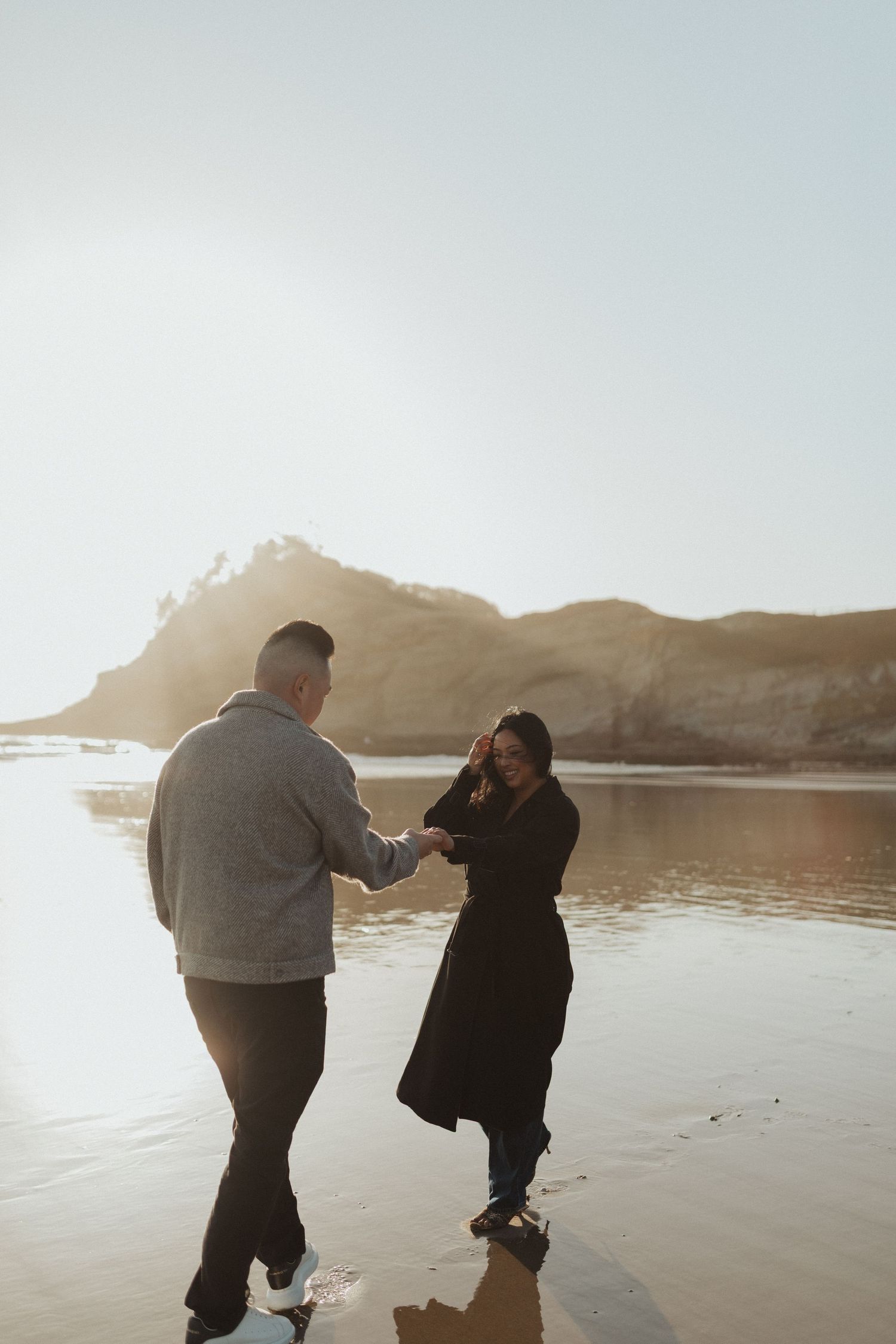 Couple holding hands and dancing on a sunlit beach with rocky cliffs in the background.