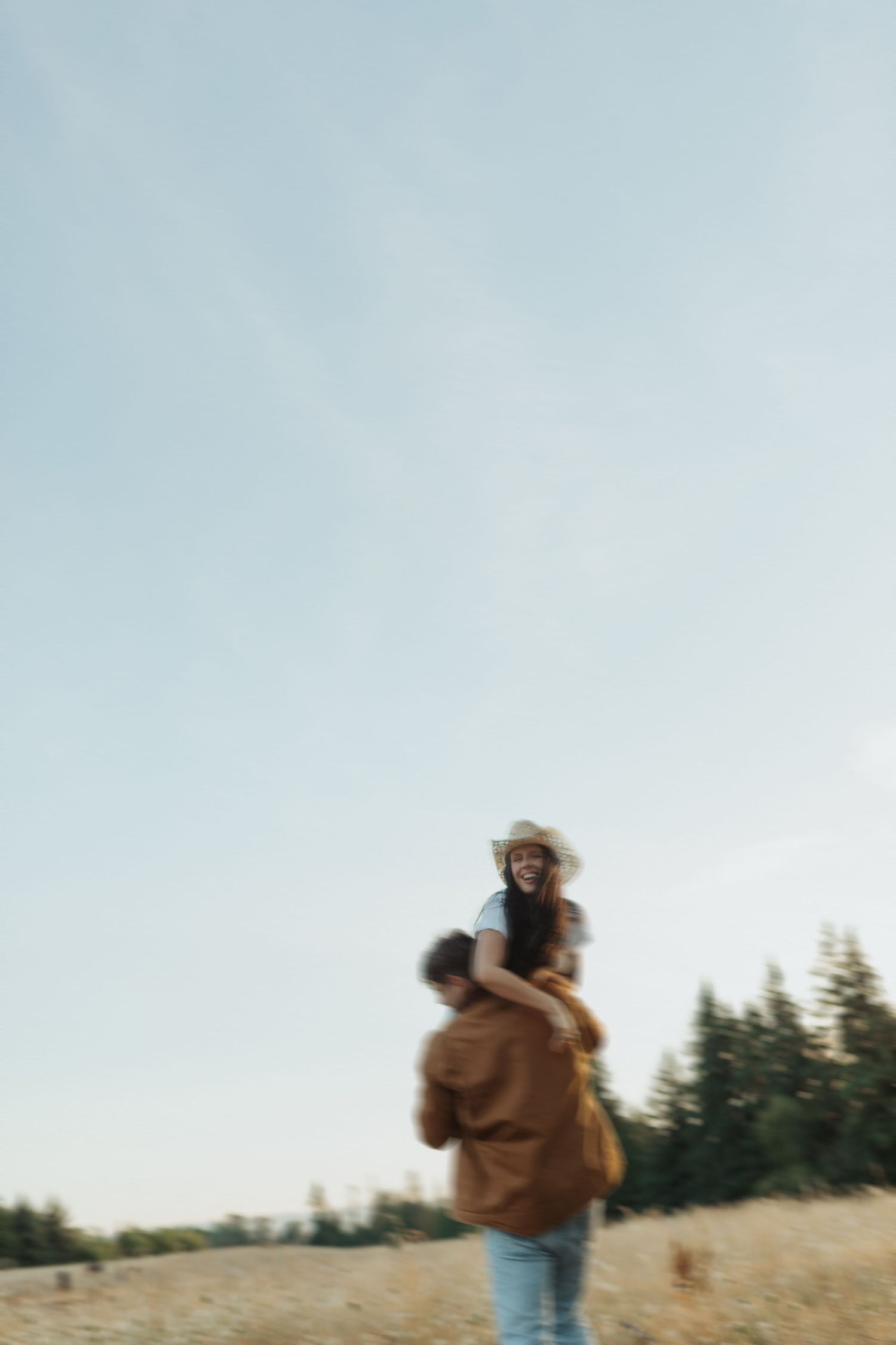 A laughing woman in a cowboy hat rides on a mans shoulders in a golden meadow with pine trees.