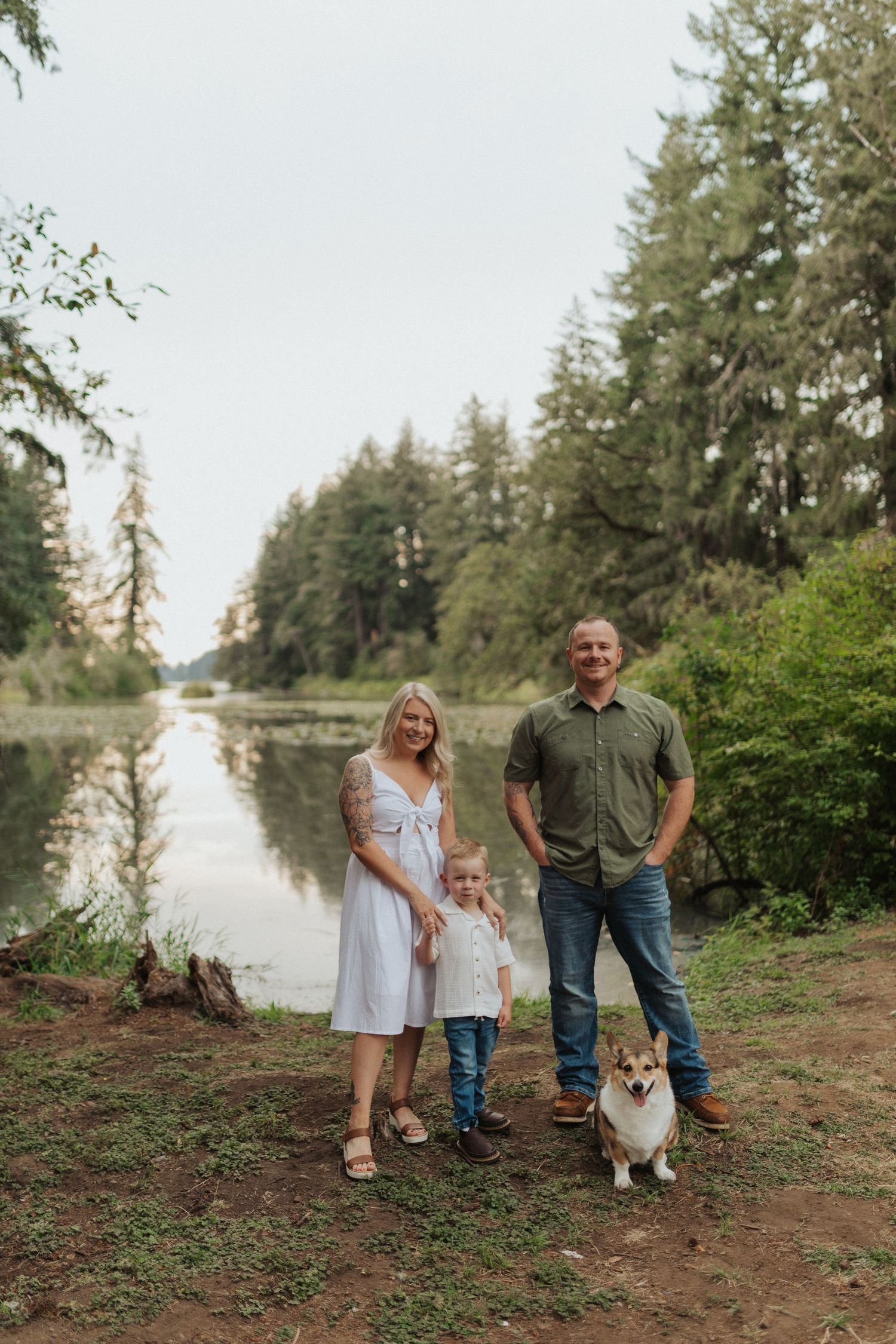 Family with young child and corgi dog posing by a peaceful tree-lined river in nature.
