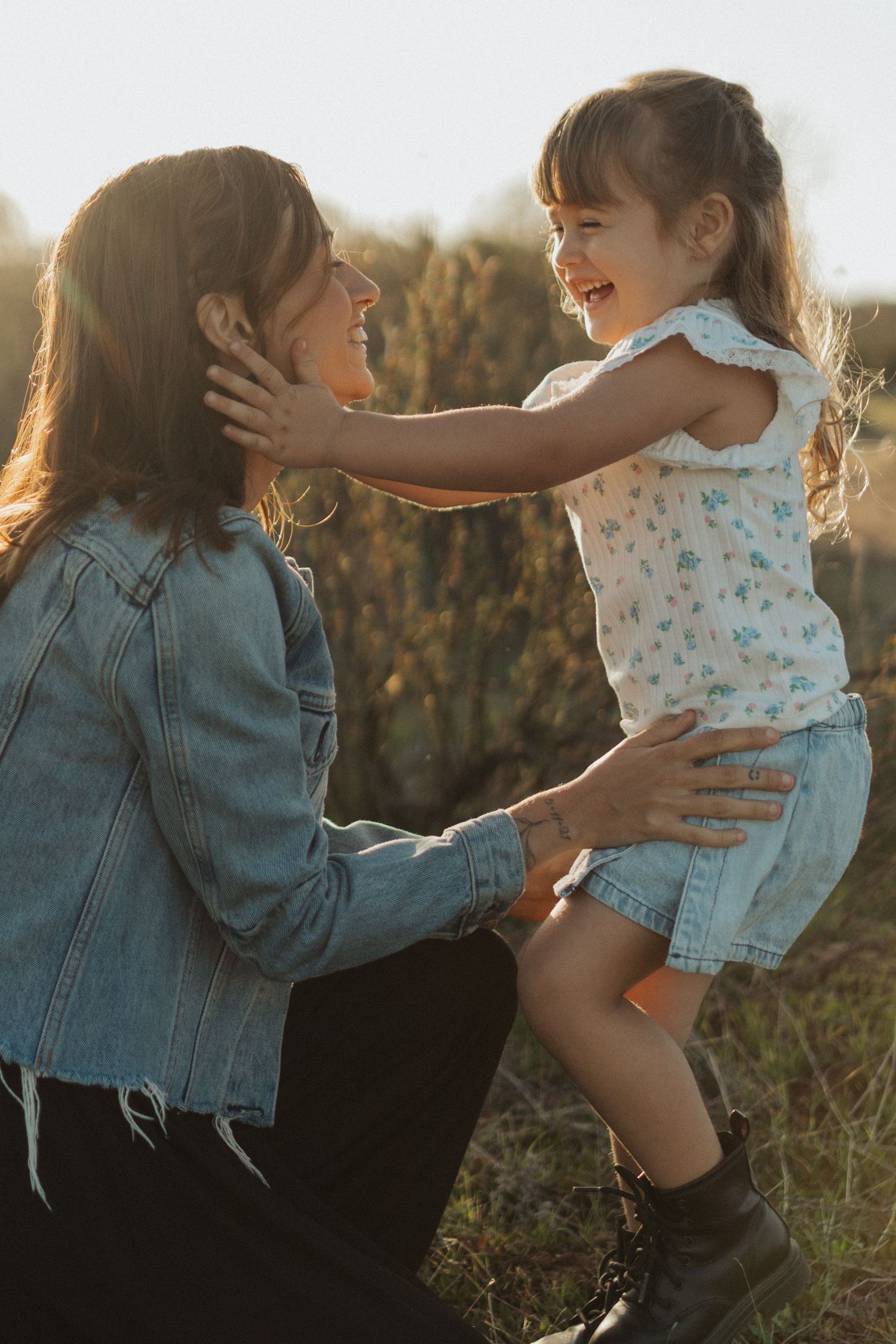 Mother in denim jacket crouching to embrace laughing young daughter in floral dress at golden hour.
