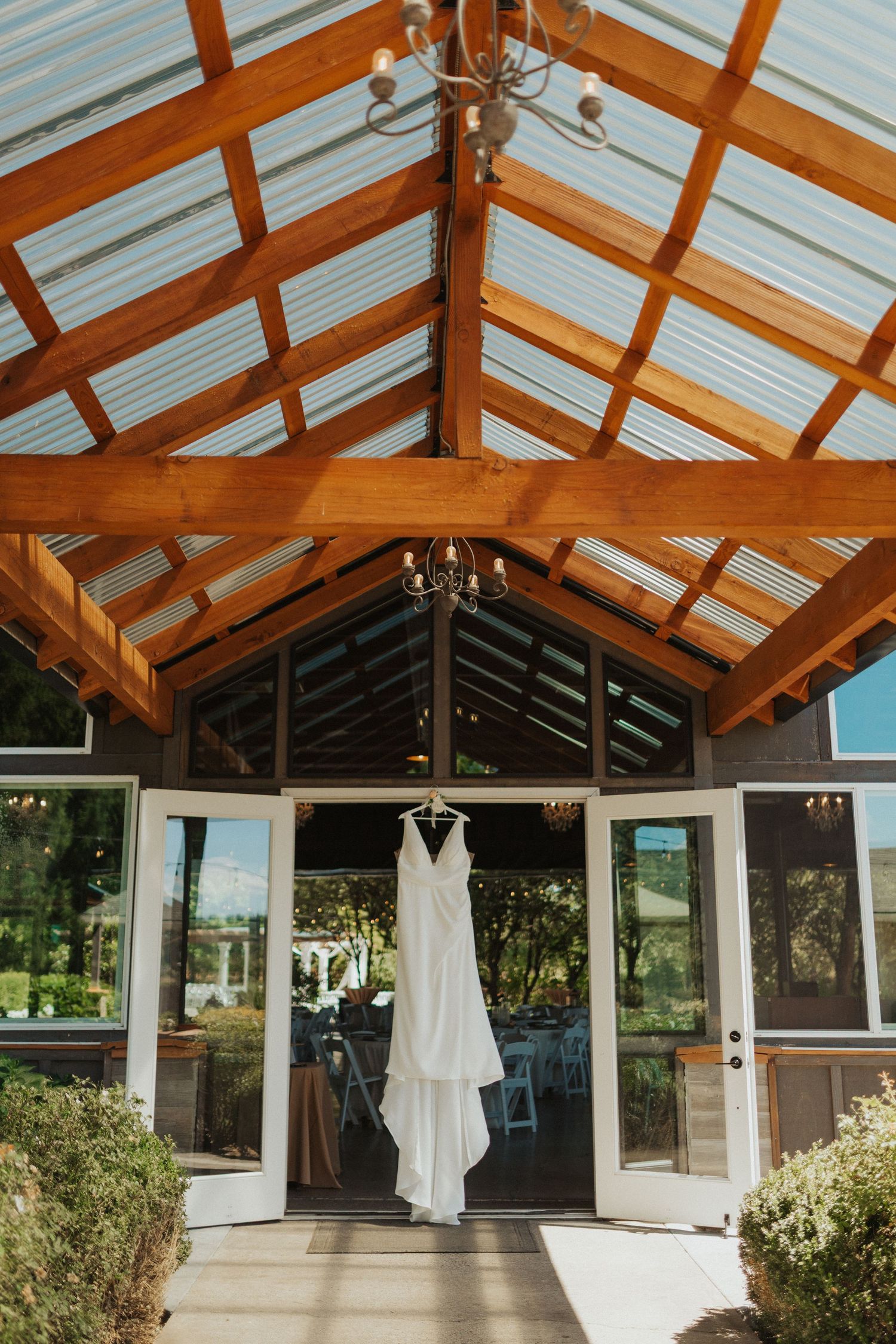 White wedding gown hanging in a rustic wooden greenhouse venue with glass ceiling and chandelier details.
